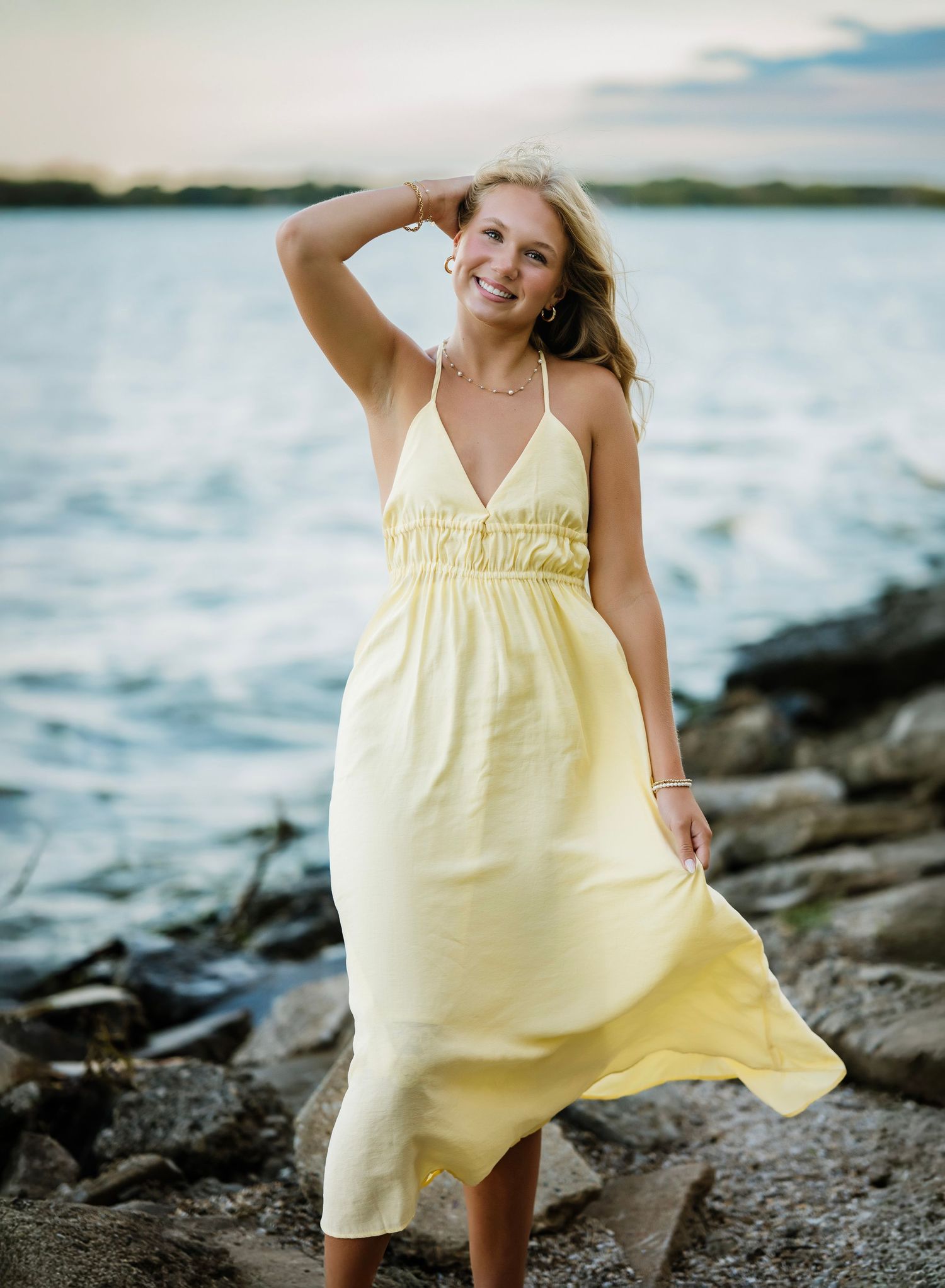 A person in a flowing yellow sundress poses playfully on a rocky beach at sunset near the ocean.