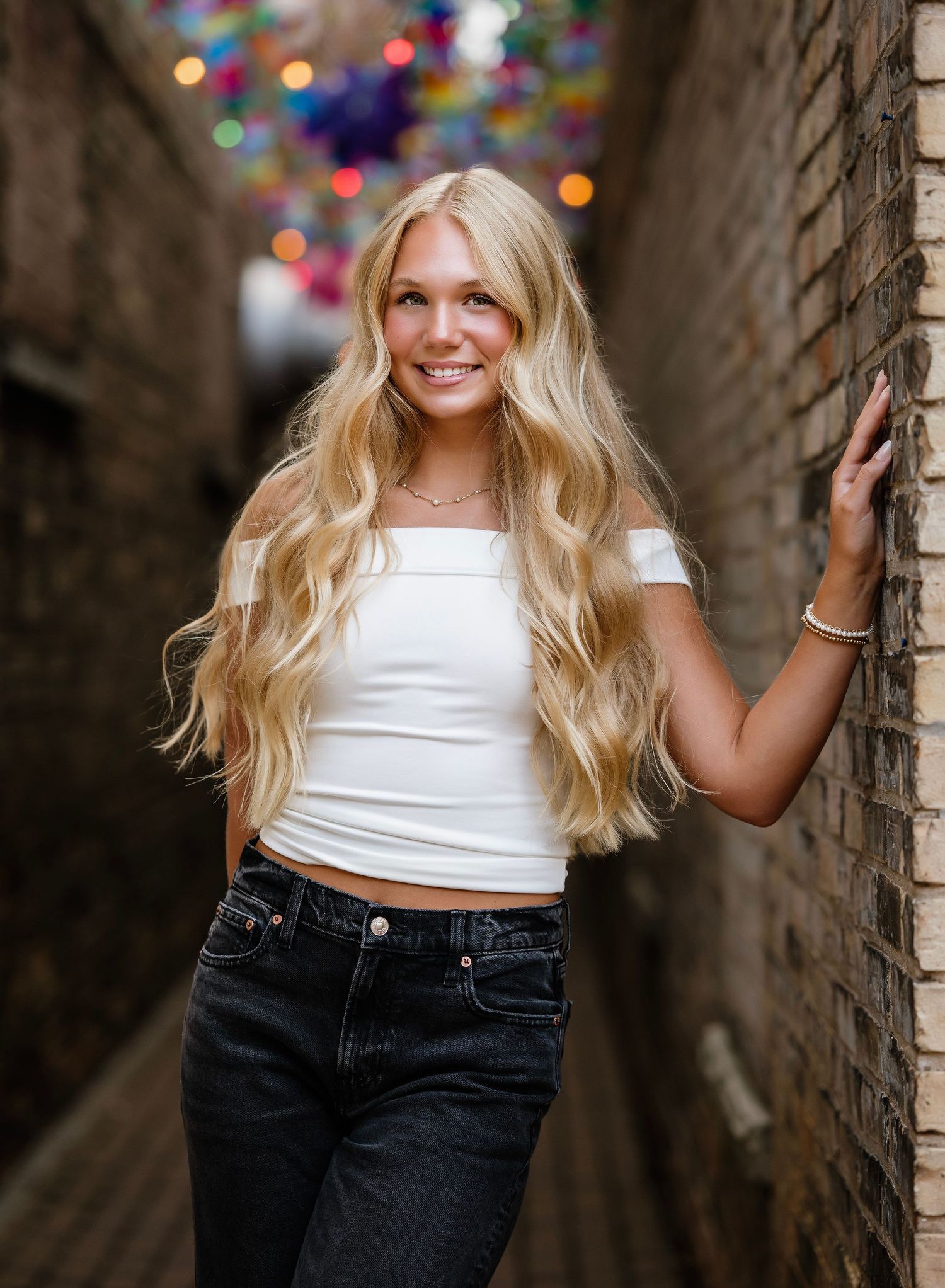 Person in a white crop top and dark jeans poses against a brick wall with colorful bokeh lights in the background.