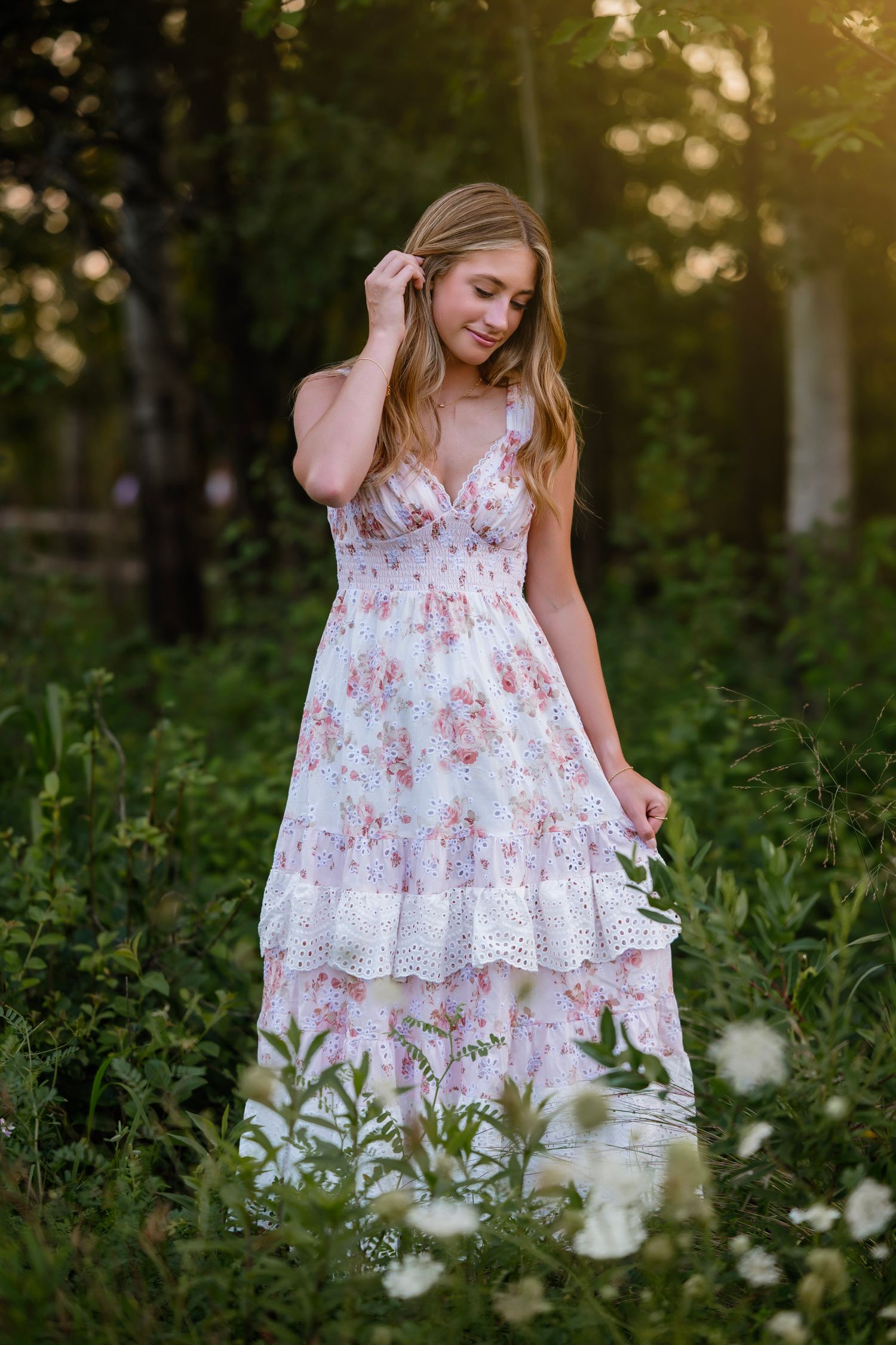 Young woman in floral maxi dress standing among wildflowers in golden hour sunlight.