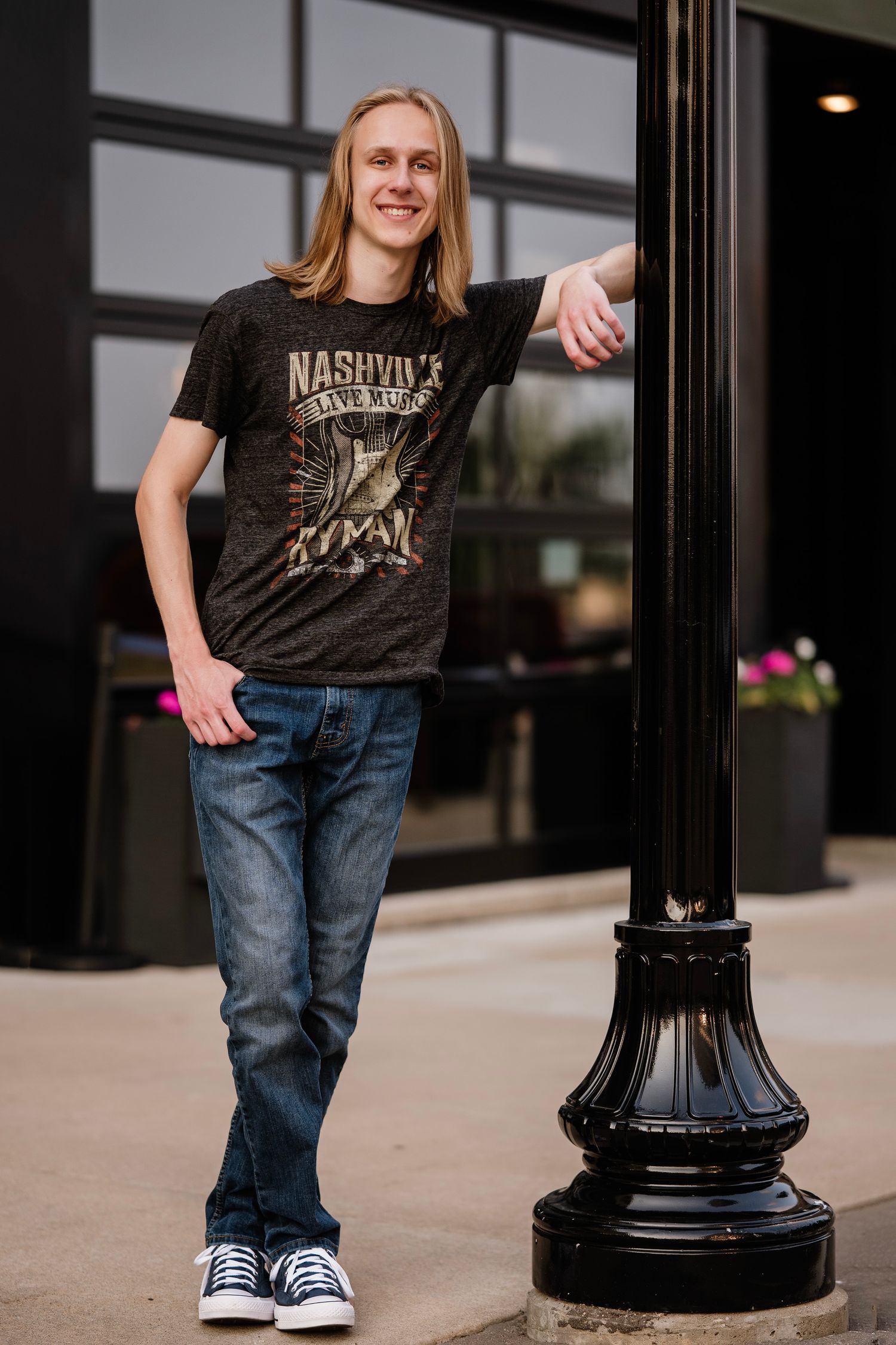 Young man with long blonde hair wearing a Nashville Live Music t-shirt and jeans, leaning on a black lamp post outdoors.