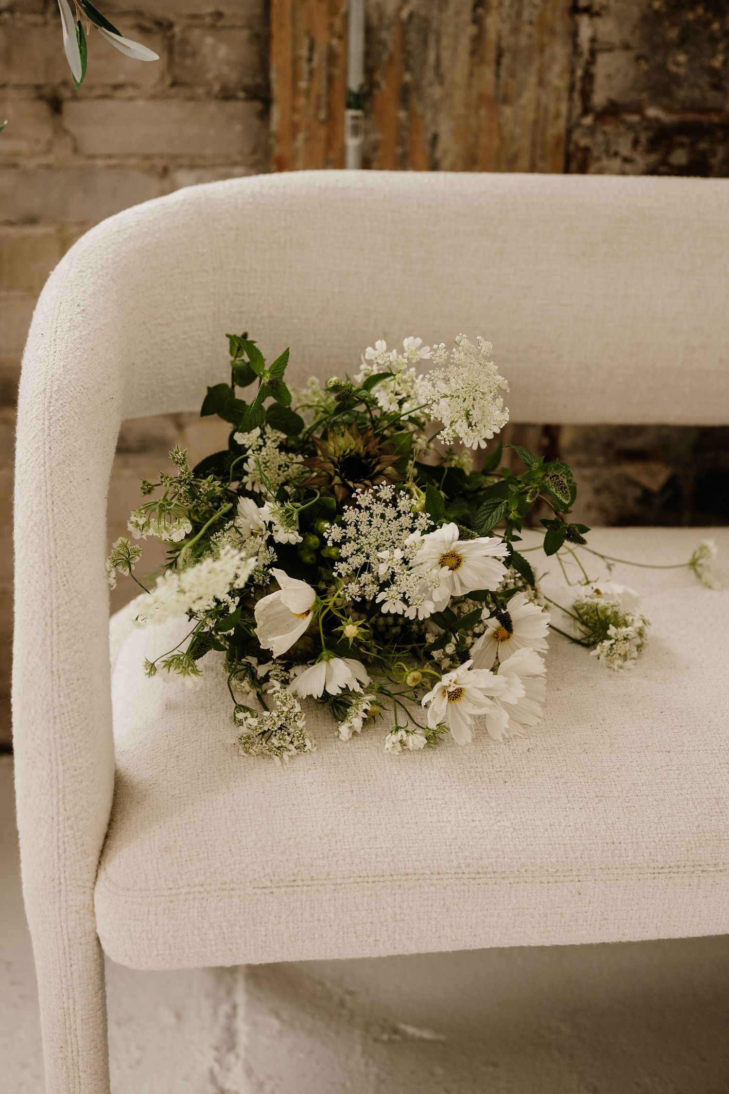 White wildflower wedding bouquet with daisies and greenery resting on a cream upholstered chair.