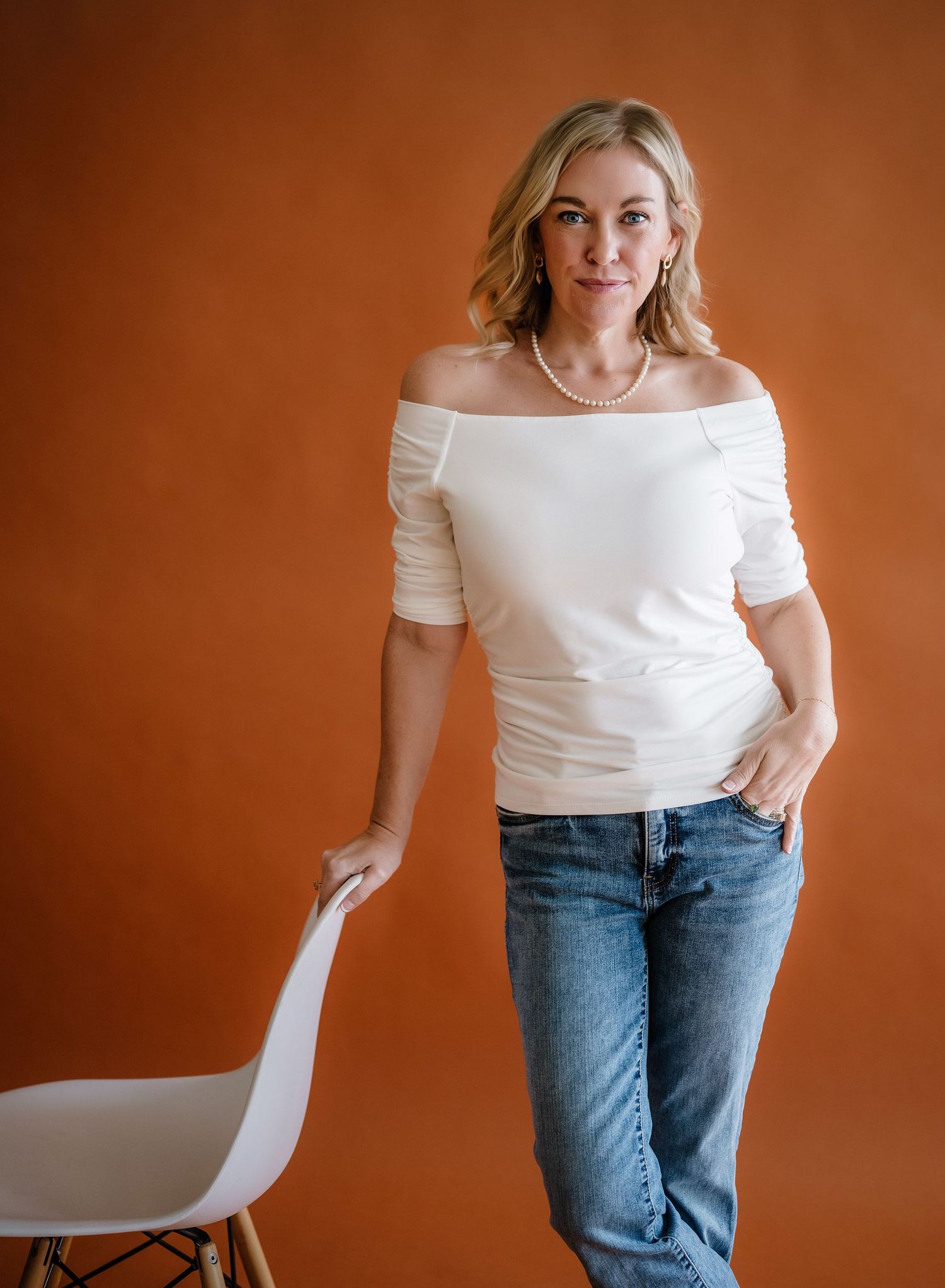 A person in a white off-shoulder top and blue jeans poses casually against an orange background with a minimalist chair.