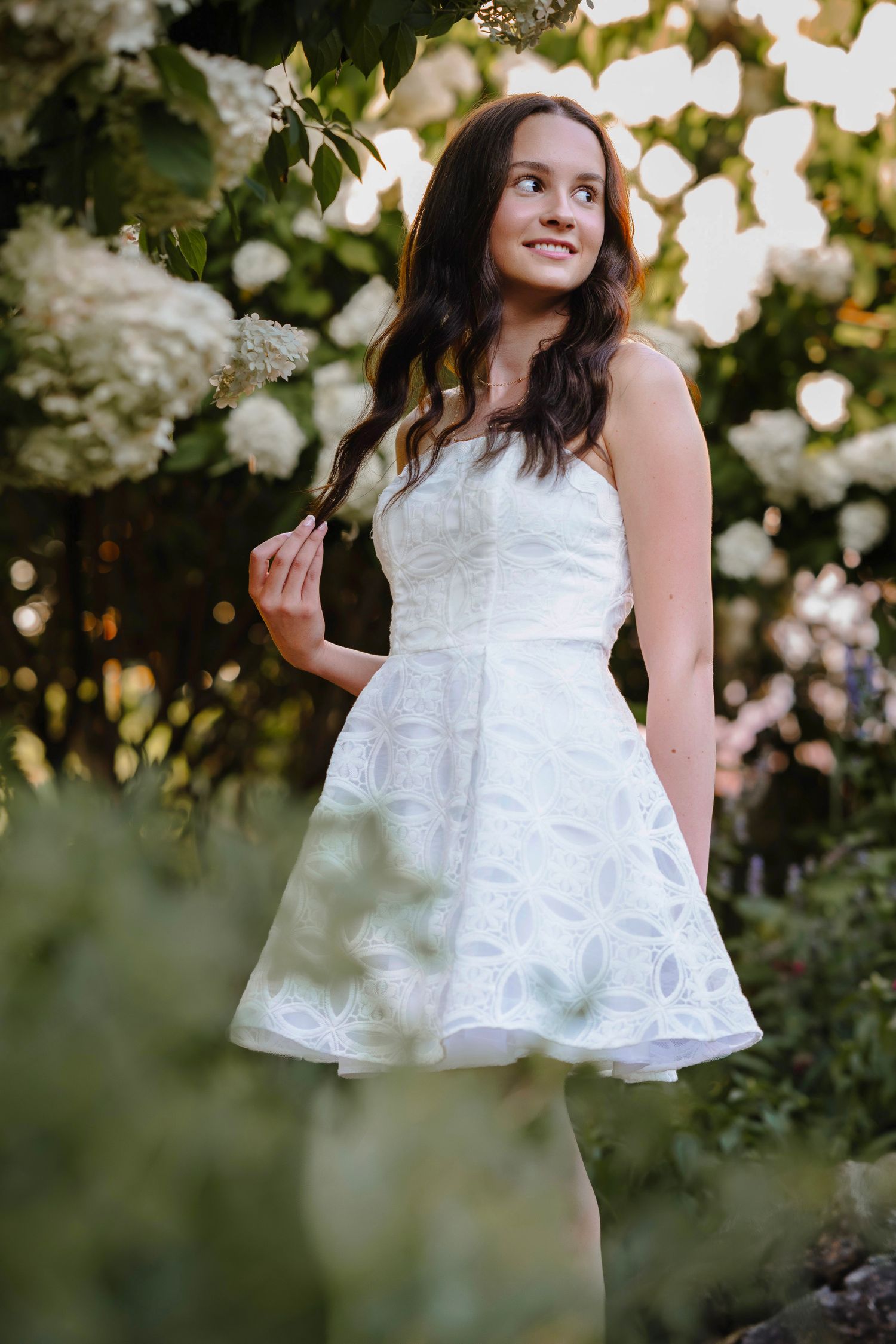 Young woman in white lace dress smiling among blooming white hydrangeas in a lush garden setting.