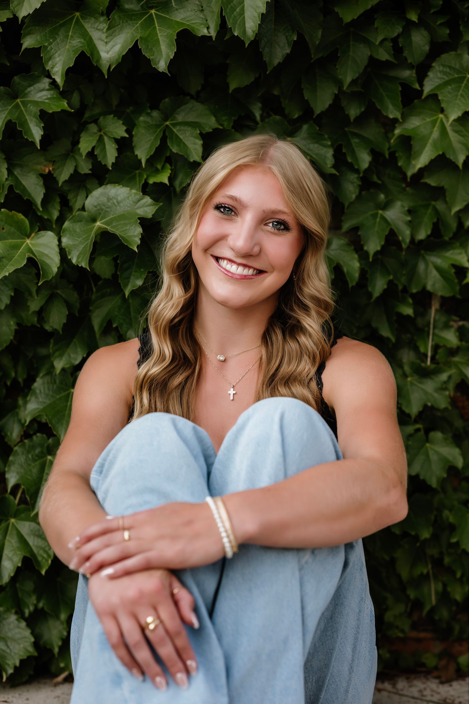 Smiling blonde woman in light blue jeans sits against a lush green ivy wall, wearing a gold cross necklace and pearl bracelet.
