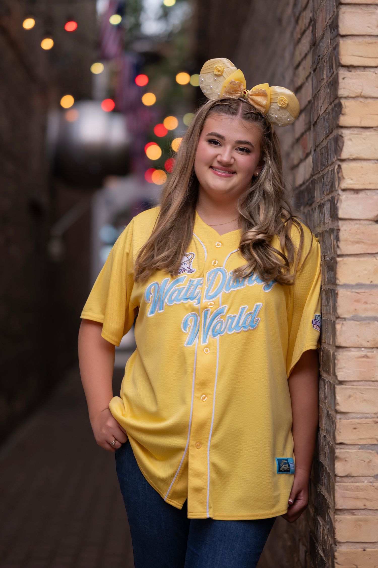Young woman in yellow Walt Disney World baseball jersey and matching Minnie Mouse ears poses against brick wall.