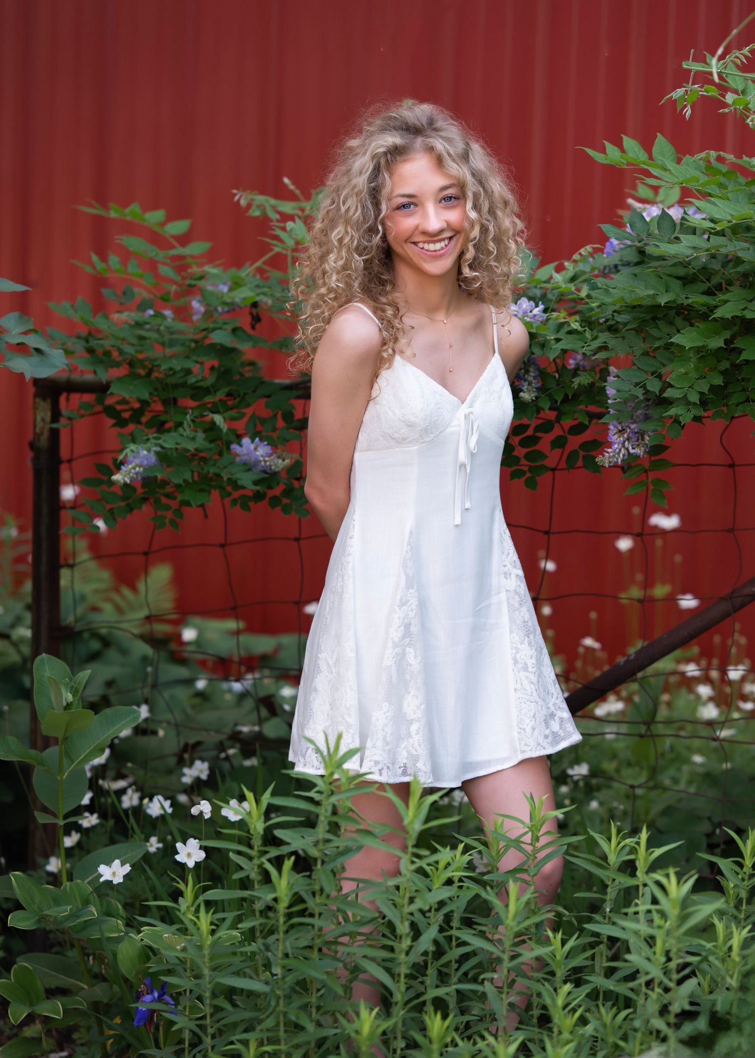 Smiling young woman with curly blonde hair wearing a white sundress, standing among green plants and flowers near a red barn.
