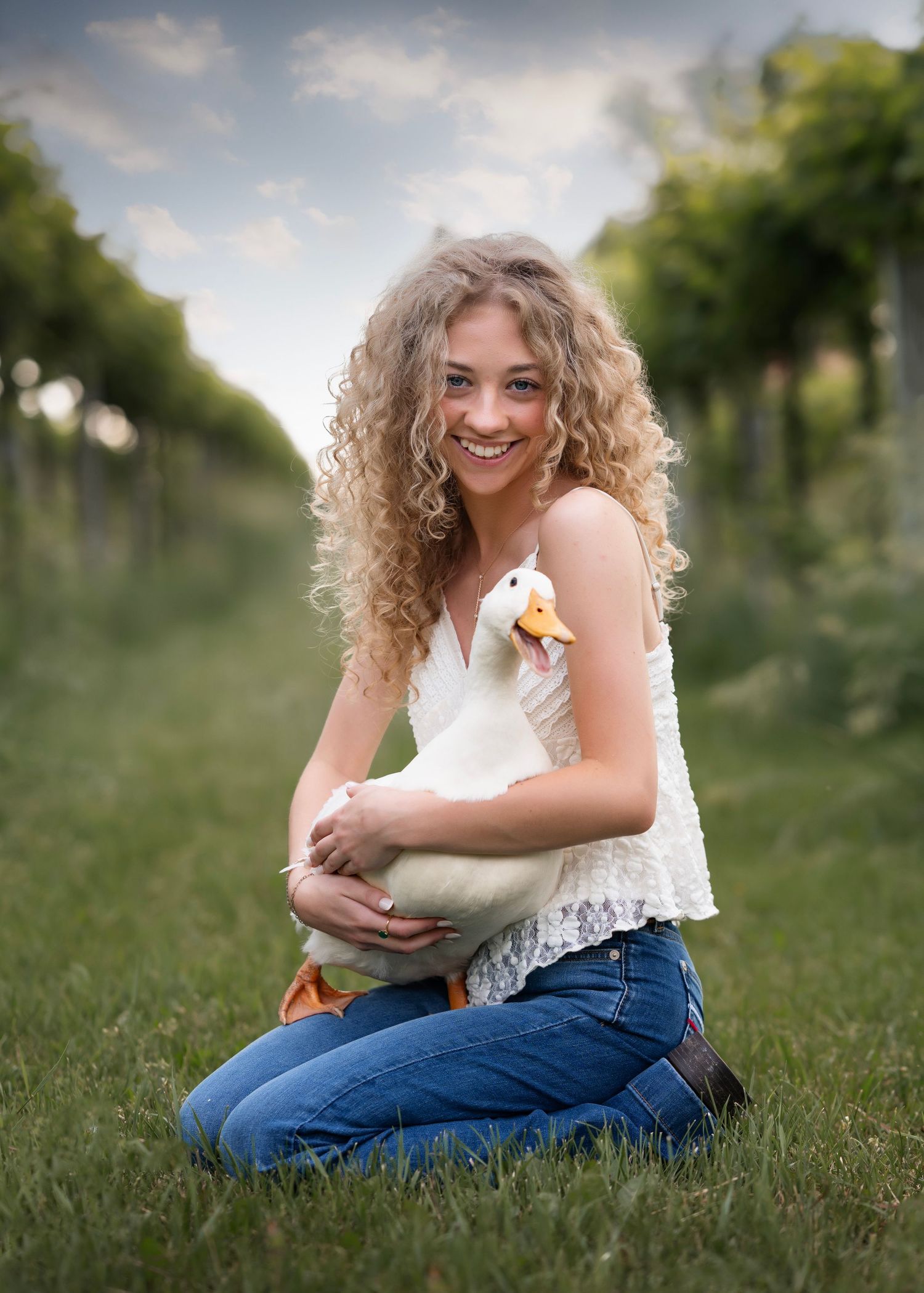 Smiling young woman with curly blonde hair holding a white duck while kneeling in a vineyard.