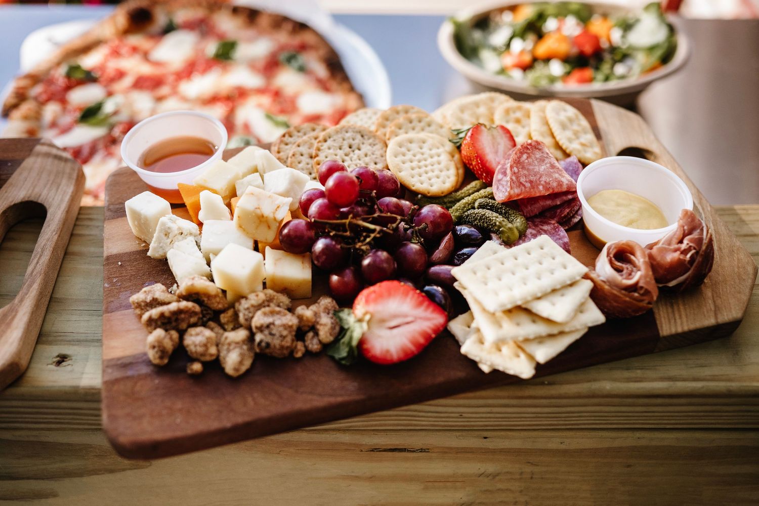 A rustic wooden charcuterie board with cheese, crackers, grapes, strawberries, walnuts, and honey beside a pizza and salad.