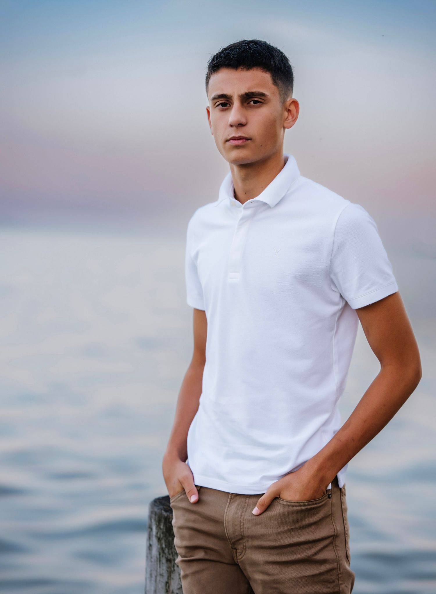 Young man in white polo shirt and khaki pants standing by the ocean at sunset with hands in pockets.