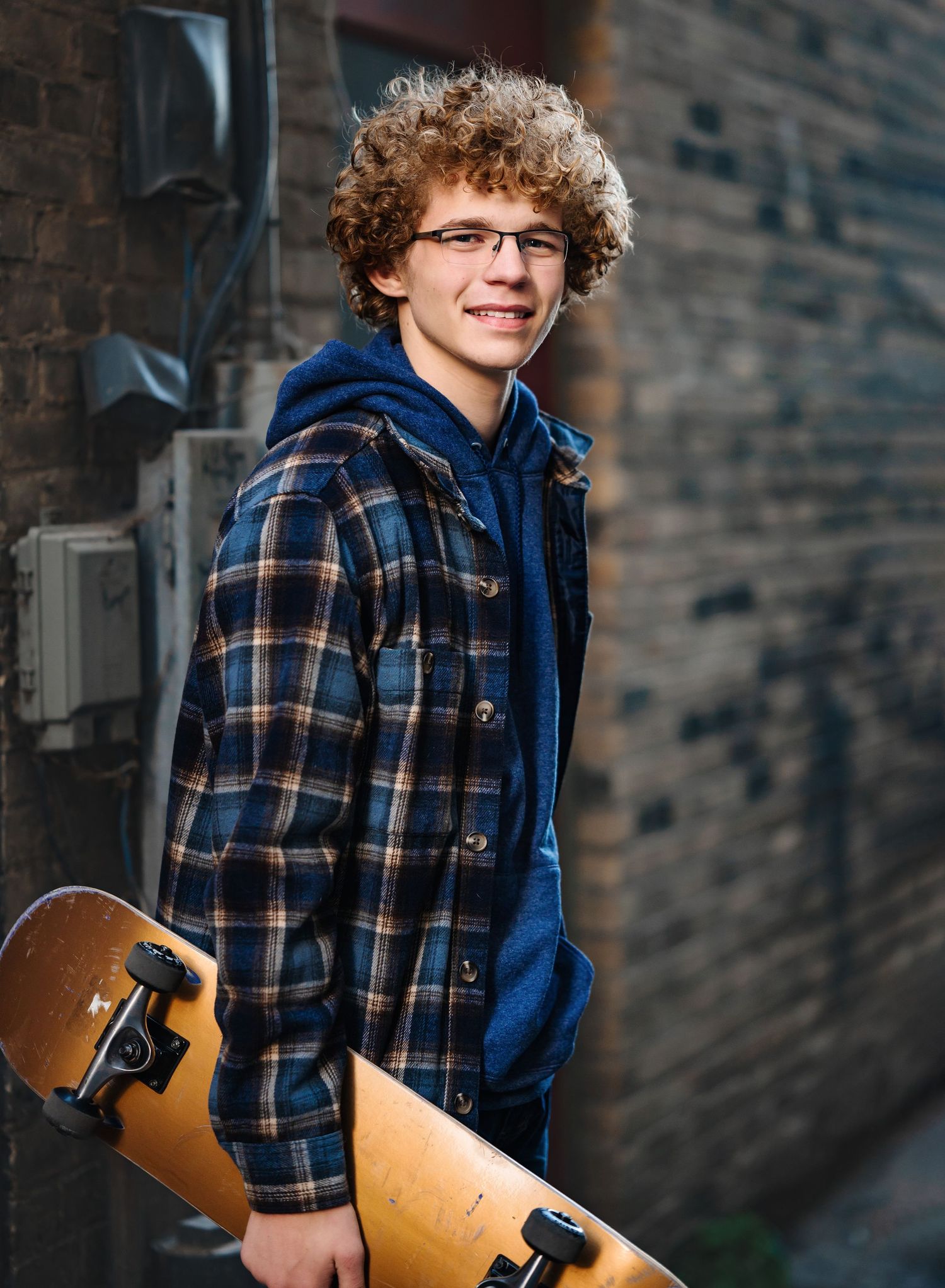 A casual urban senior portrait with plaid jacket and skateboard against industrial brick wall.