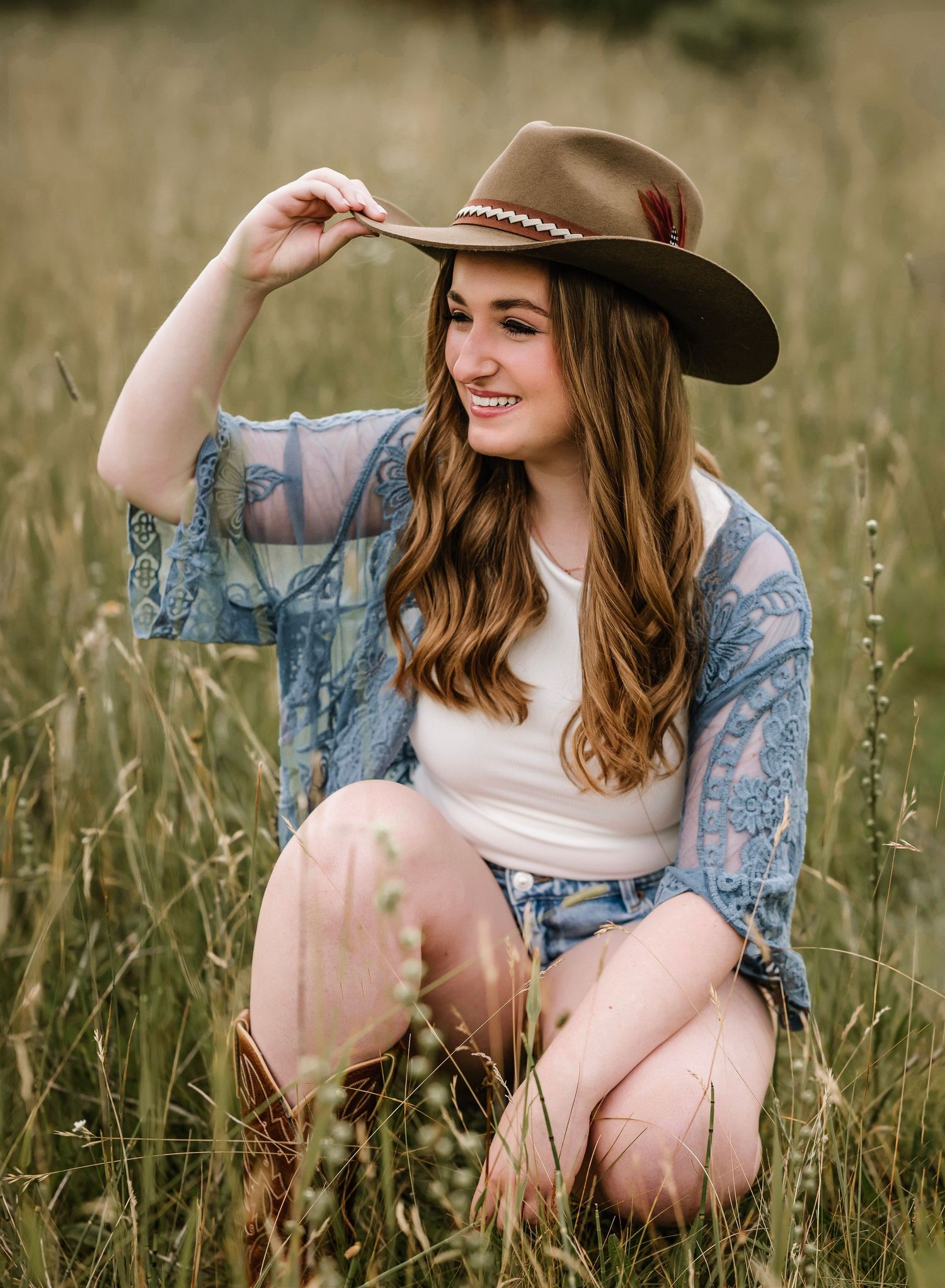 A woman in a brown cowboy hat and denim jacket sits in a tall grass field with a joyful expression.