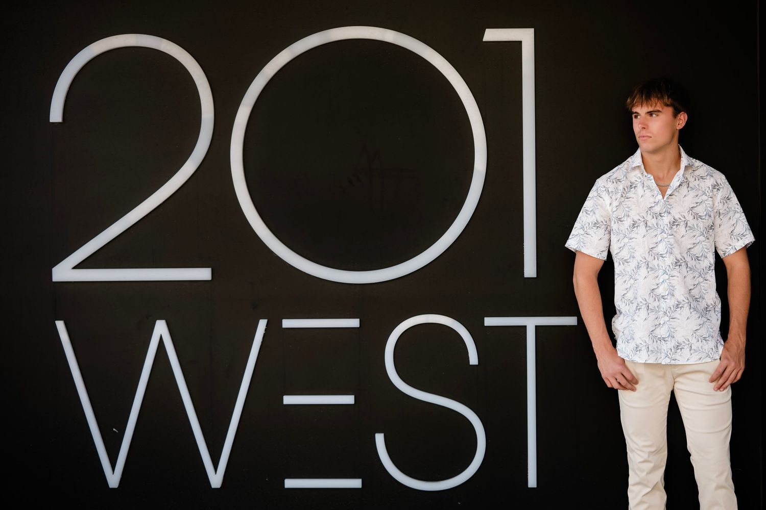 Person wearing a white patterned shirt stands against a dark wall with '201 WEST' text displayed.
