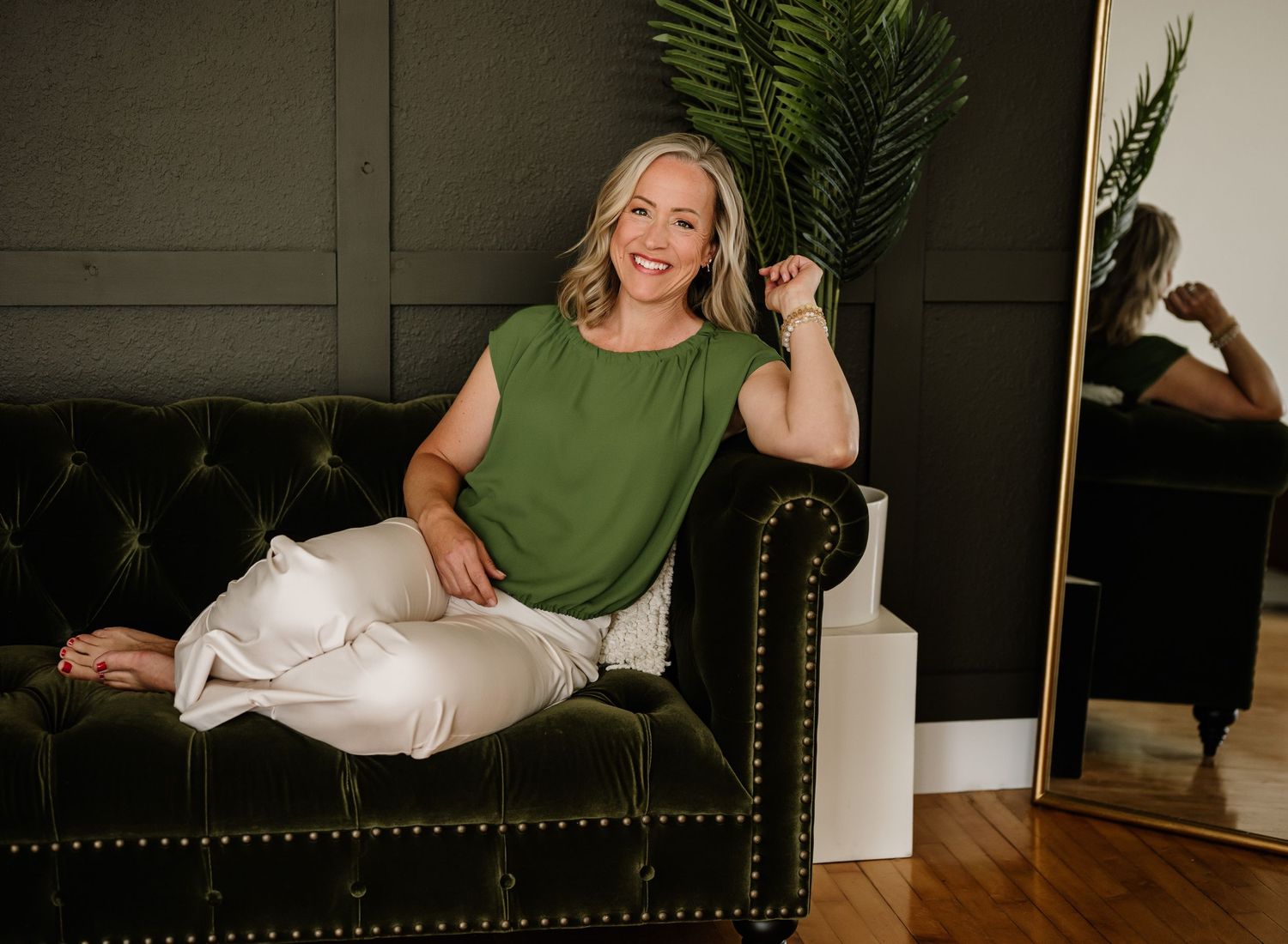 Woman in a green sleeveless top and white pants sits casually on a dark leather chair next to a potted palm plant.