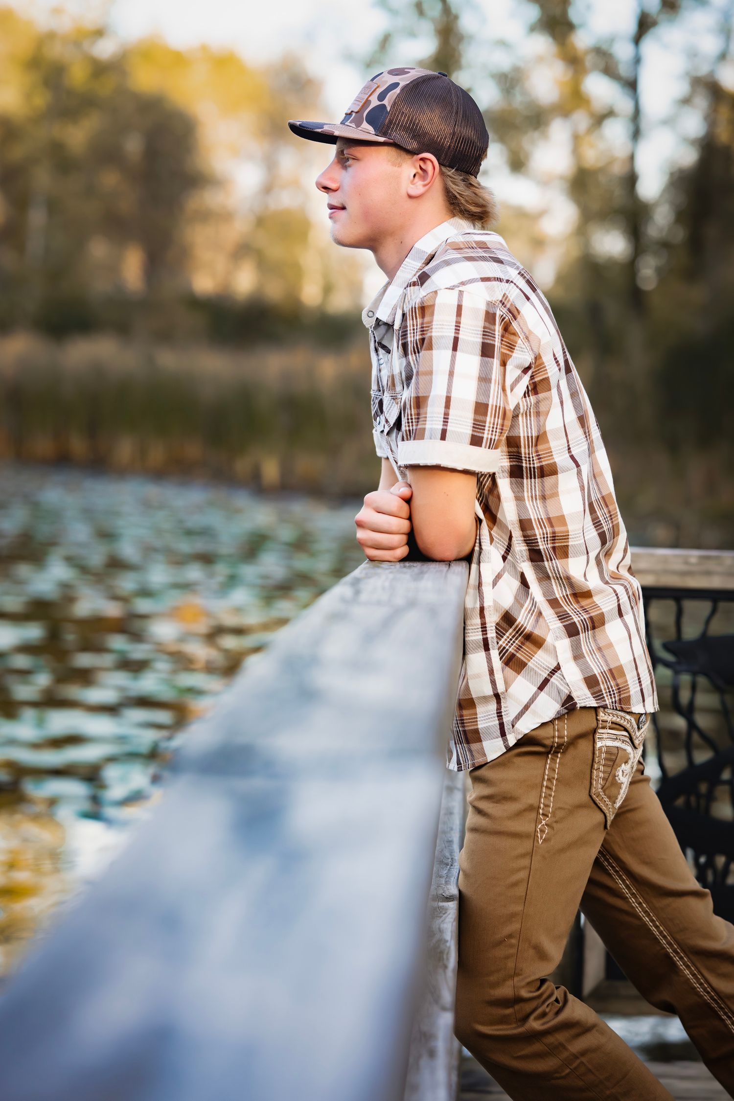 A person in a plaid shirt and brown pants sits casually on a wooden dock by a serene lake with lily pads.