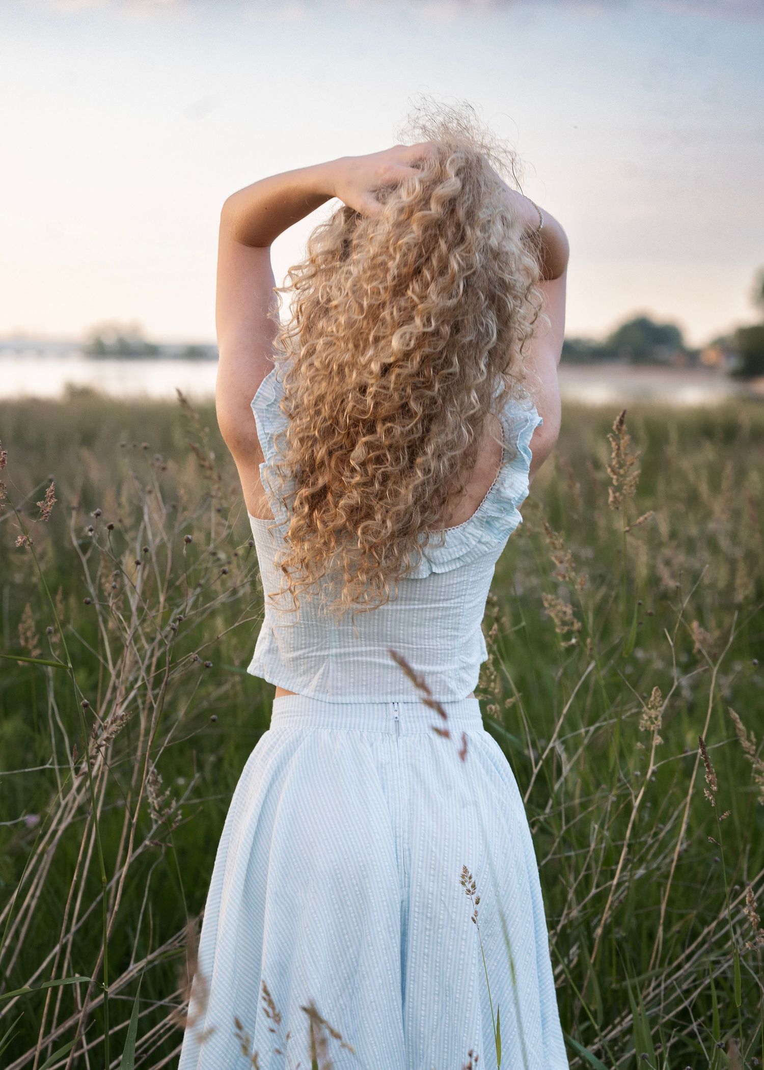 Woman with long curly blonde hair in a light blue dress standing in a field at dusk.