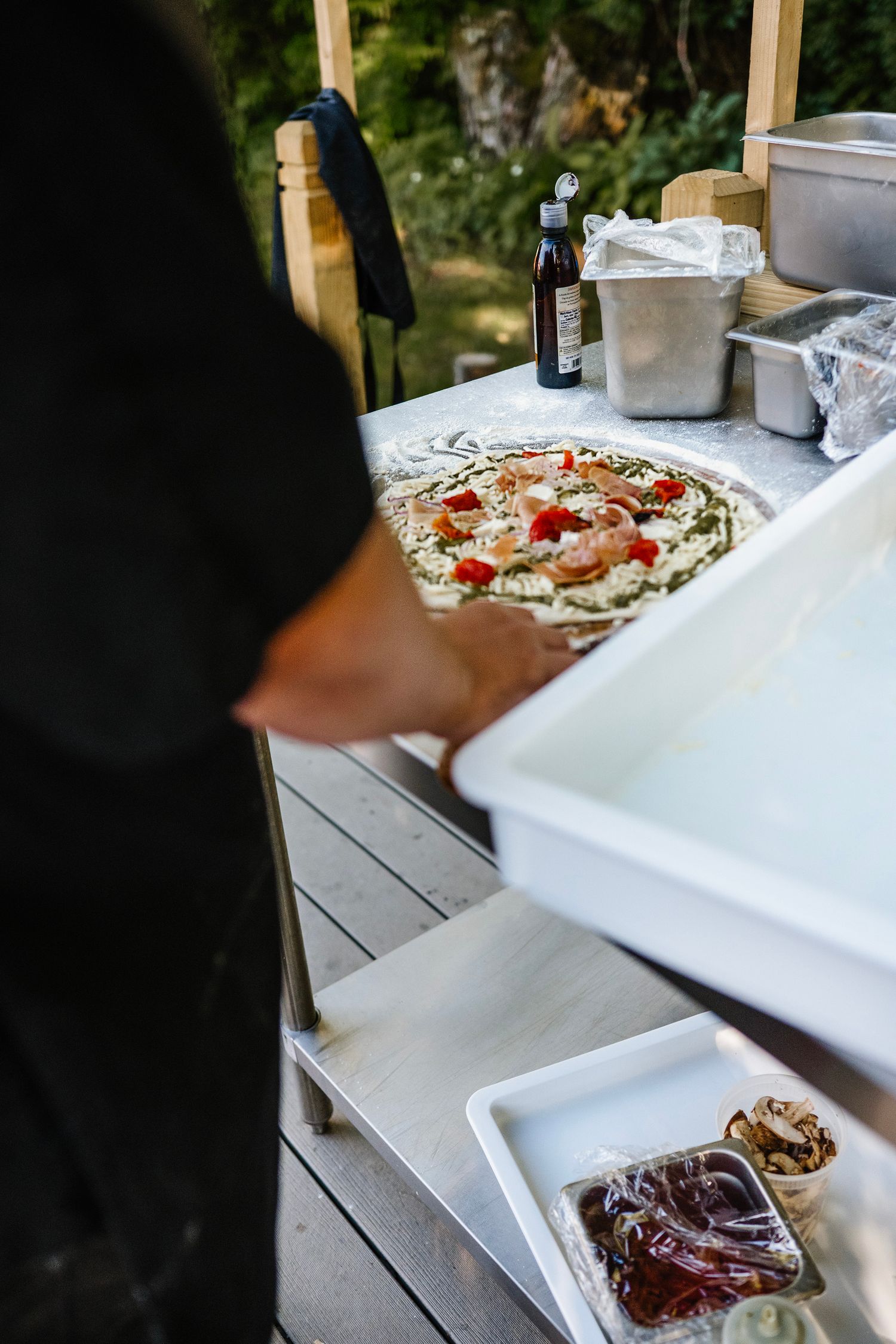 Chef preparing a homemade pizza with fresh toppings on an outdoor wooden prep station.