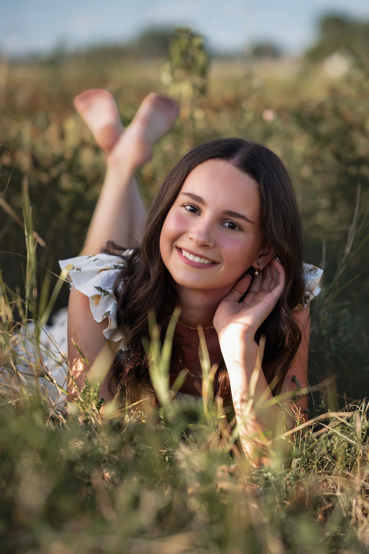 Young woman with long brown hair lying in a grassy field, smiling warmly at camera in golden hour light.