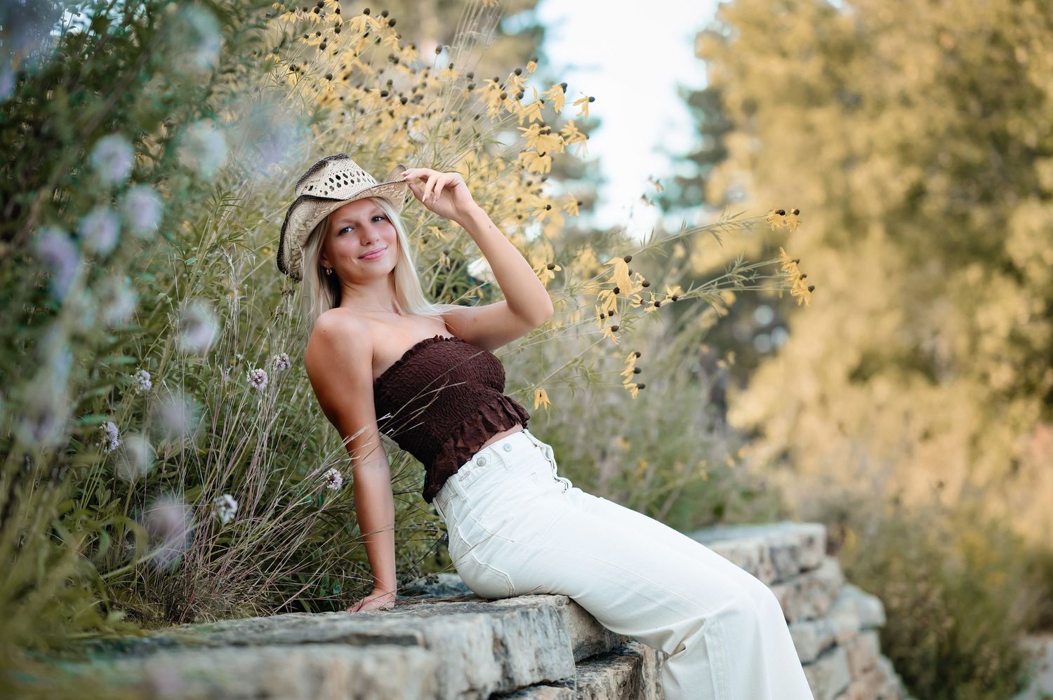 Young blonde woman in white pants and brown top poses on stone wall wearing cowboy hat among wildflowers.