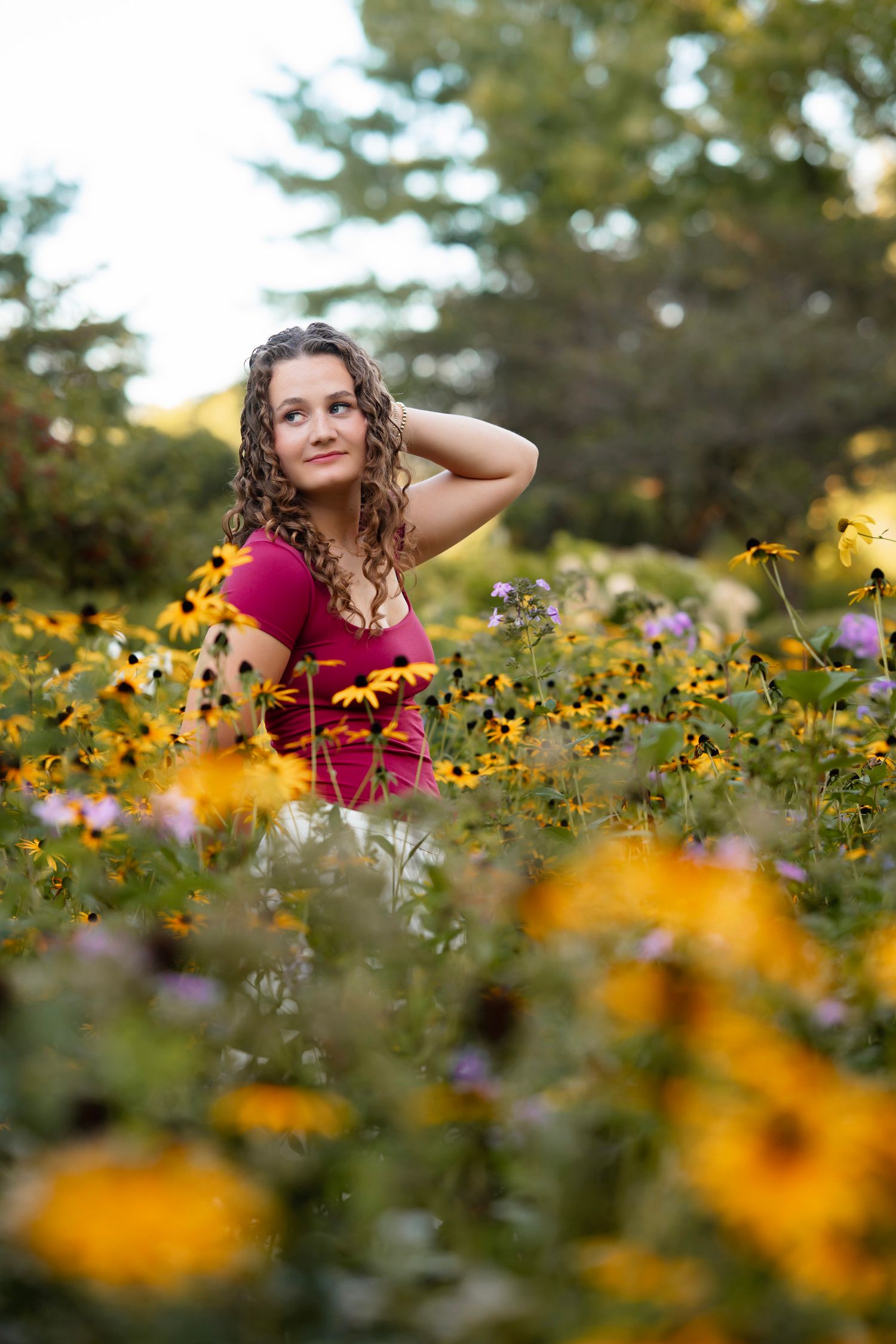 A person in a burgundy dress poses among vibrant yellow wildflowers in a summer meadow during golden hour.