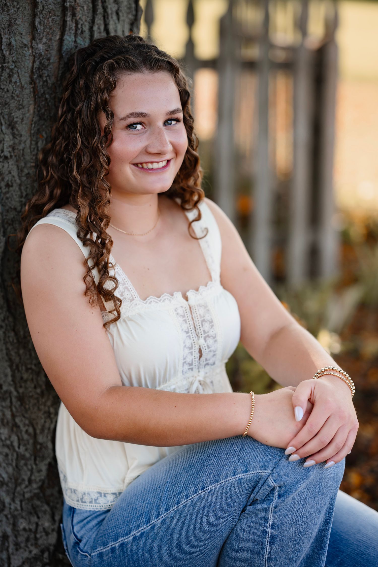A person in a white lace top and jeans sits casually against a wooden fence during golden hour.