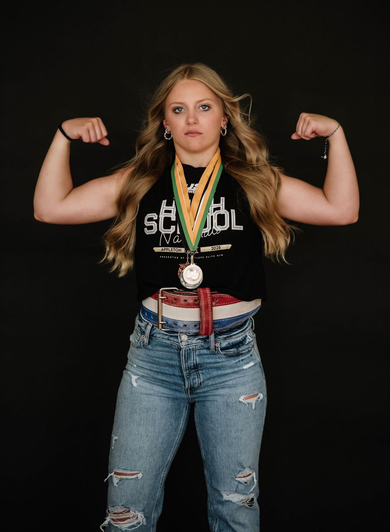 Student athlete in ripped jeans and varsity jacket shows off medals and poses with strength against dark background.