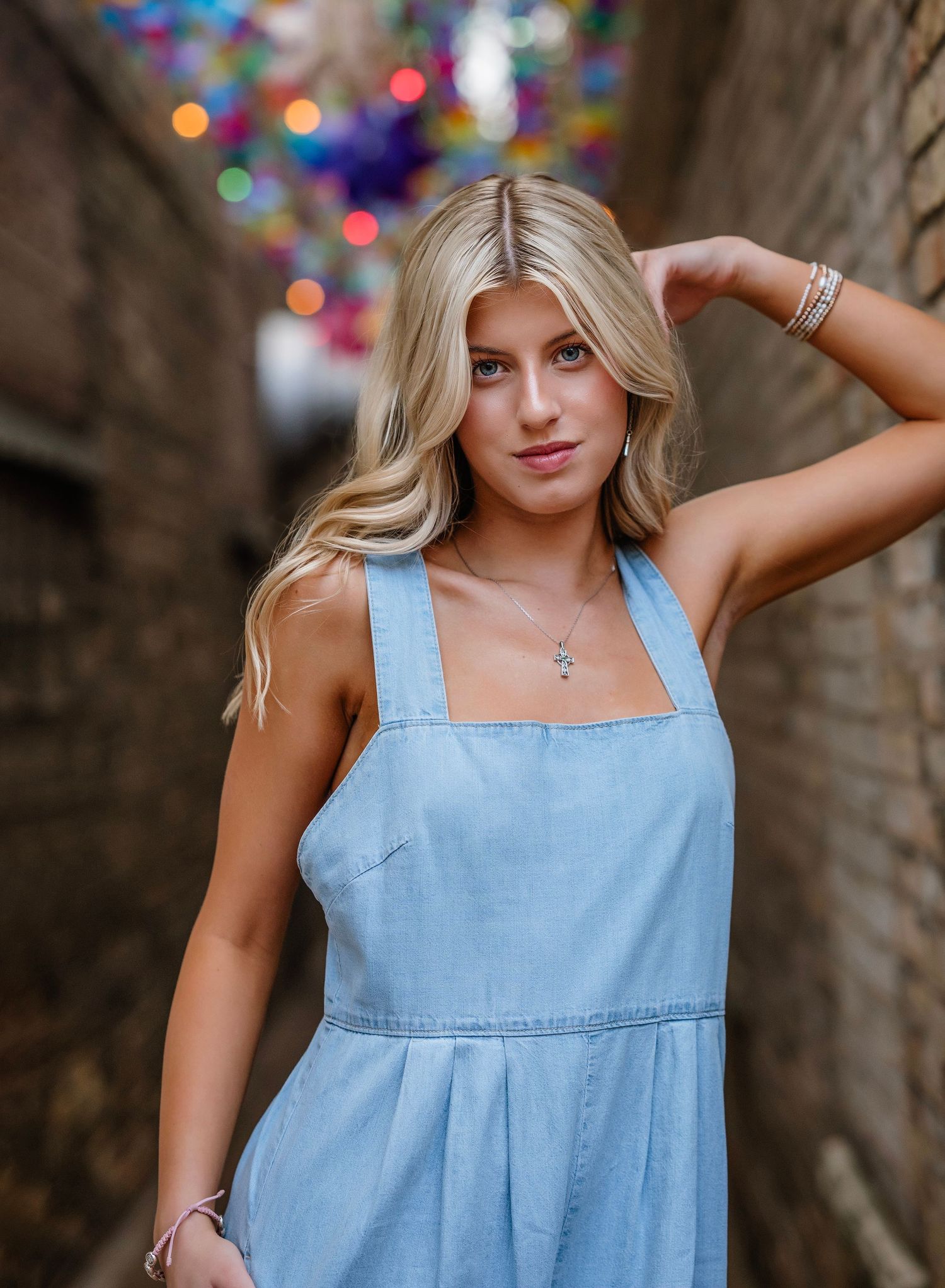 Woman in light blue denim sundress poses outdoors against blurred colorful bokeh background.