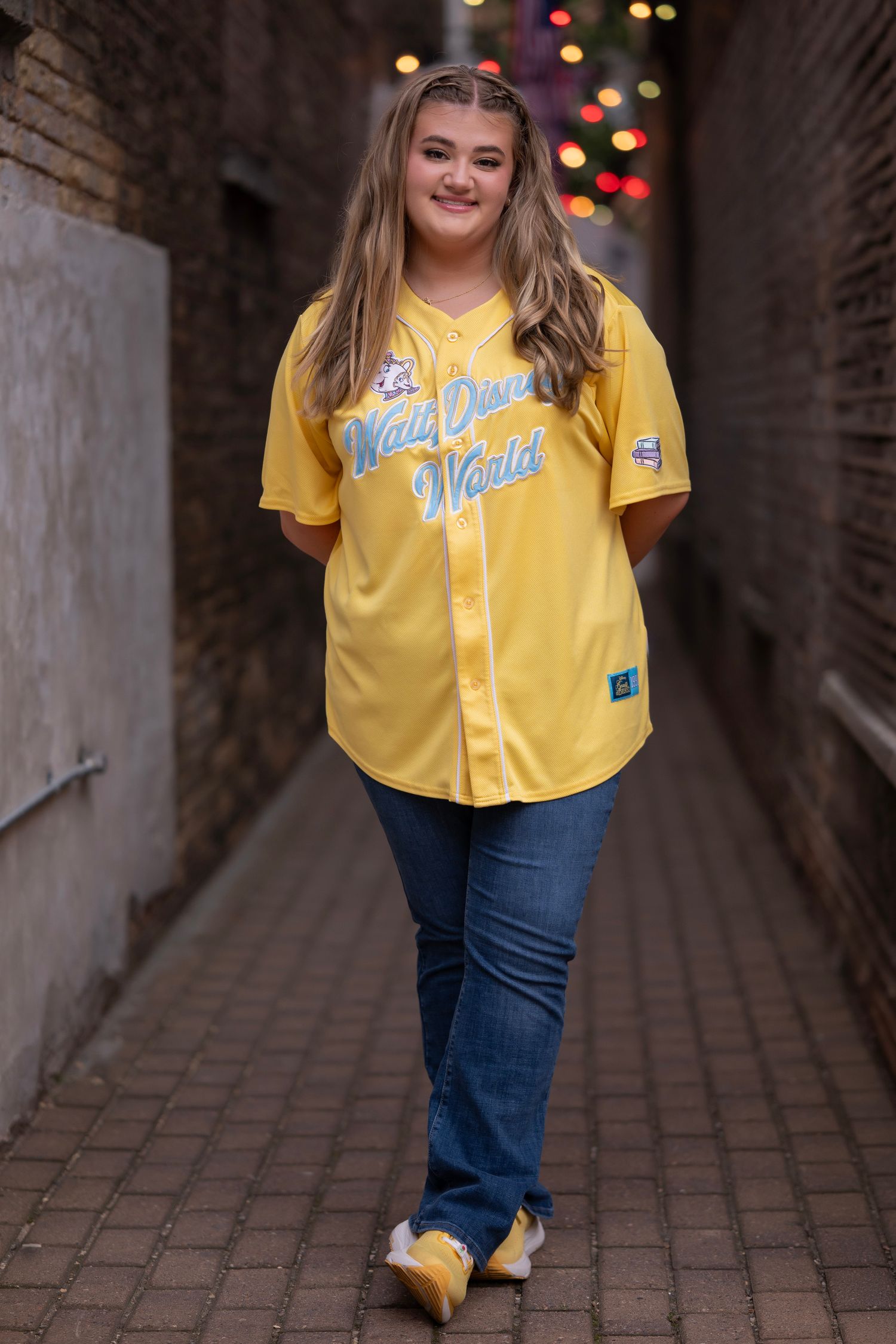 Young woman smiling in yellow Walt Disney World baseball jersey and jeans, posing in a brick alleyway with bokeh lights.