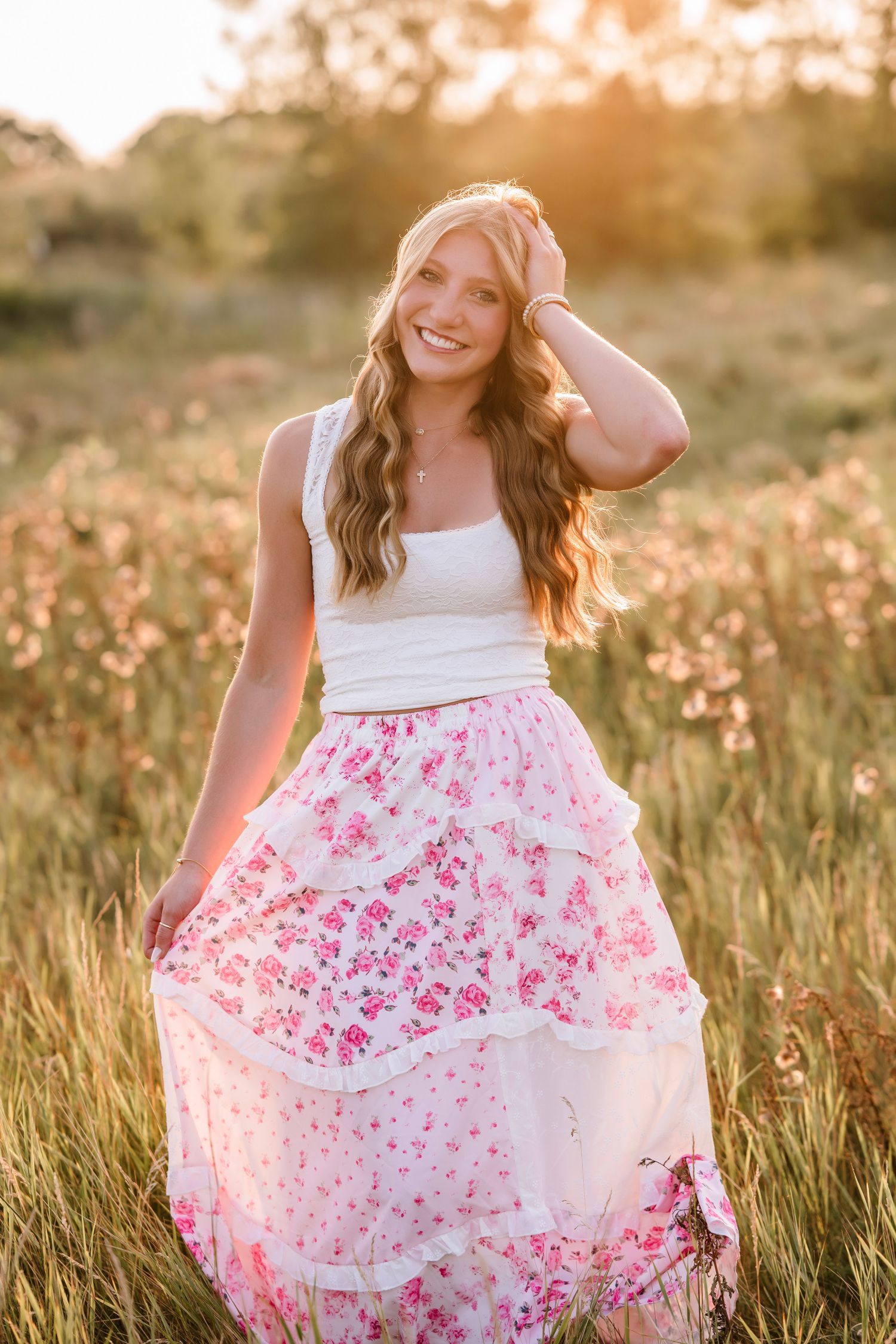 Young woman in white crop top and pink floral maxi skirt posing in golden hour sunlit meadow.
