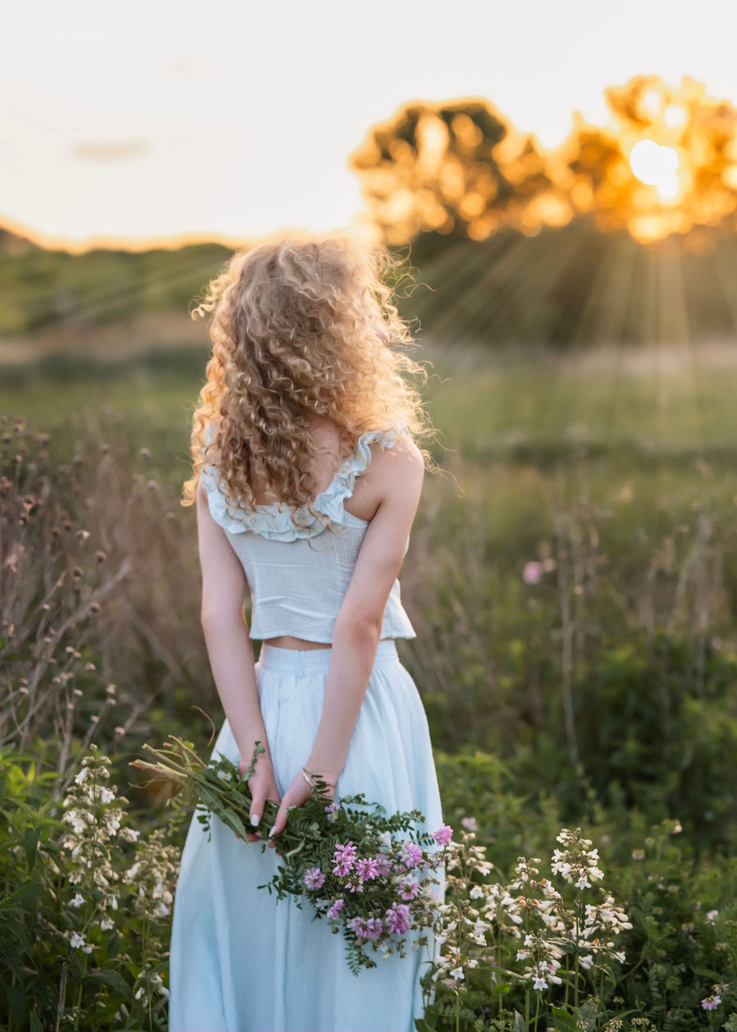 A young woman with curly hair holds wildflowers in a sunlit meadow, wearing a white crop top and skirt at golden hour.