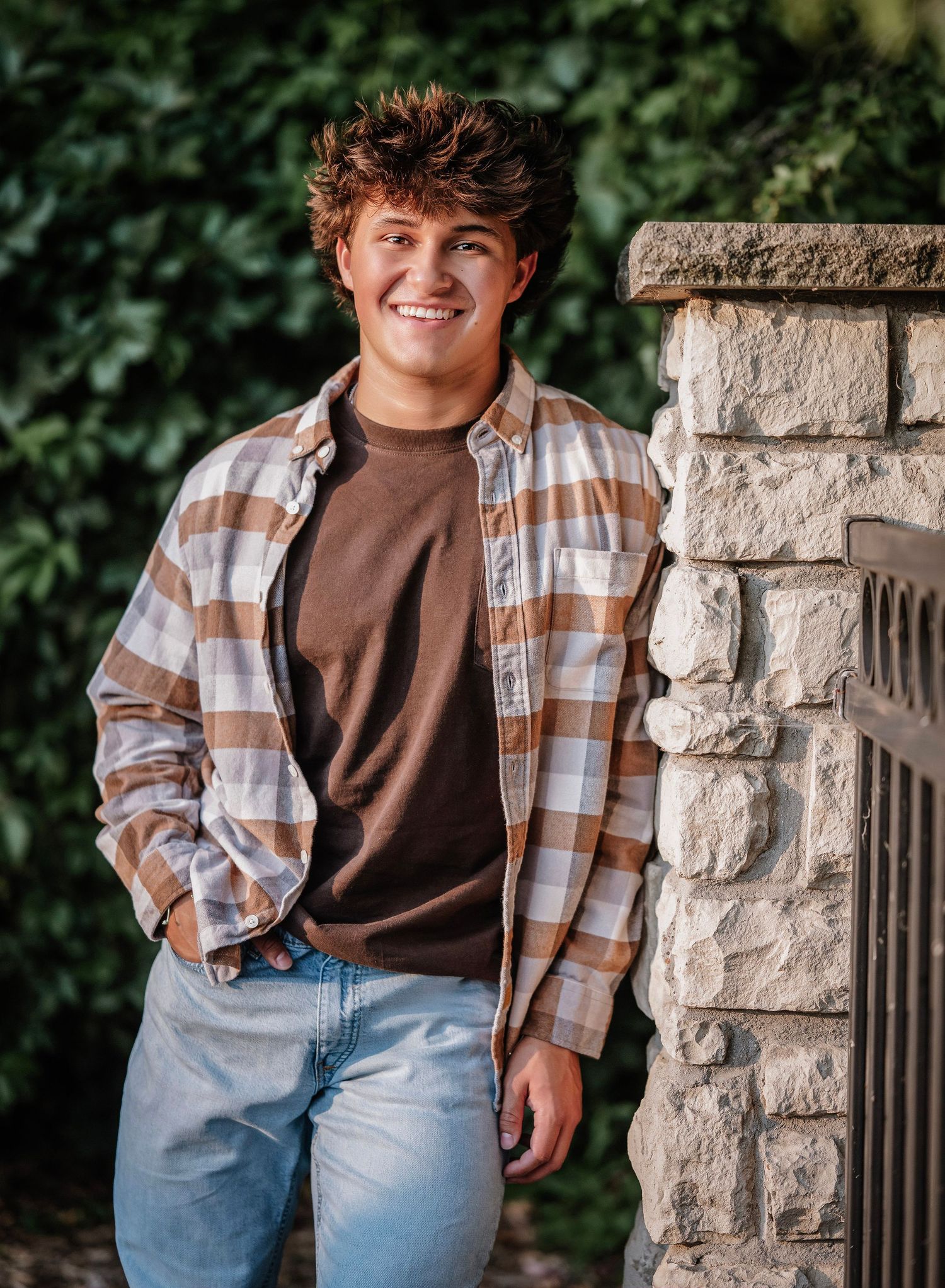 Person wearing a plaid flannel shirt and jeans poses casually against a stone pillar in an outdoor setting.