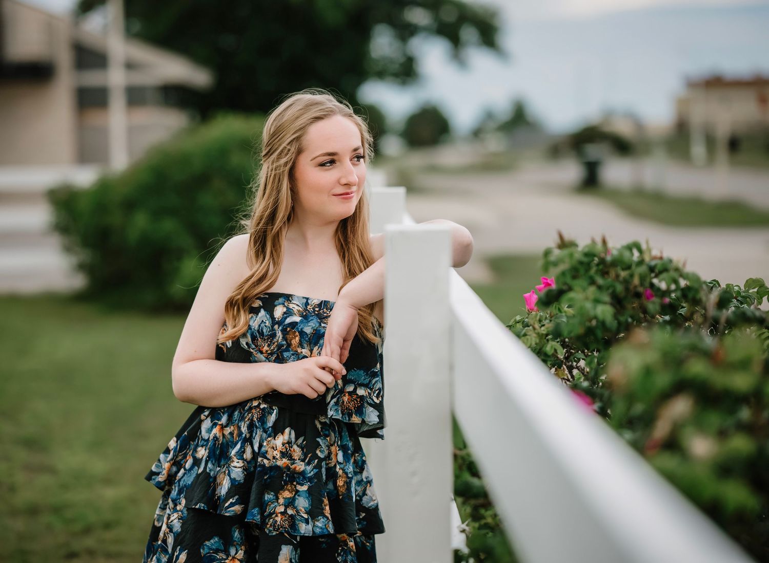 A person in a floral dress poses gracefully against a white railing in an outdoor setting.