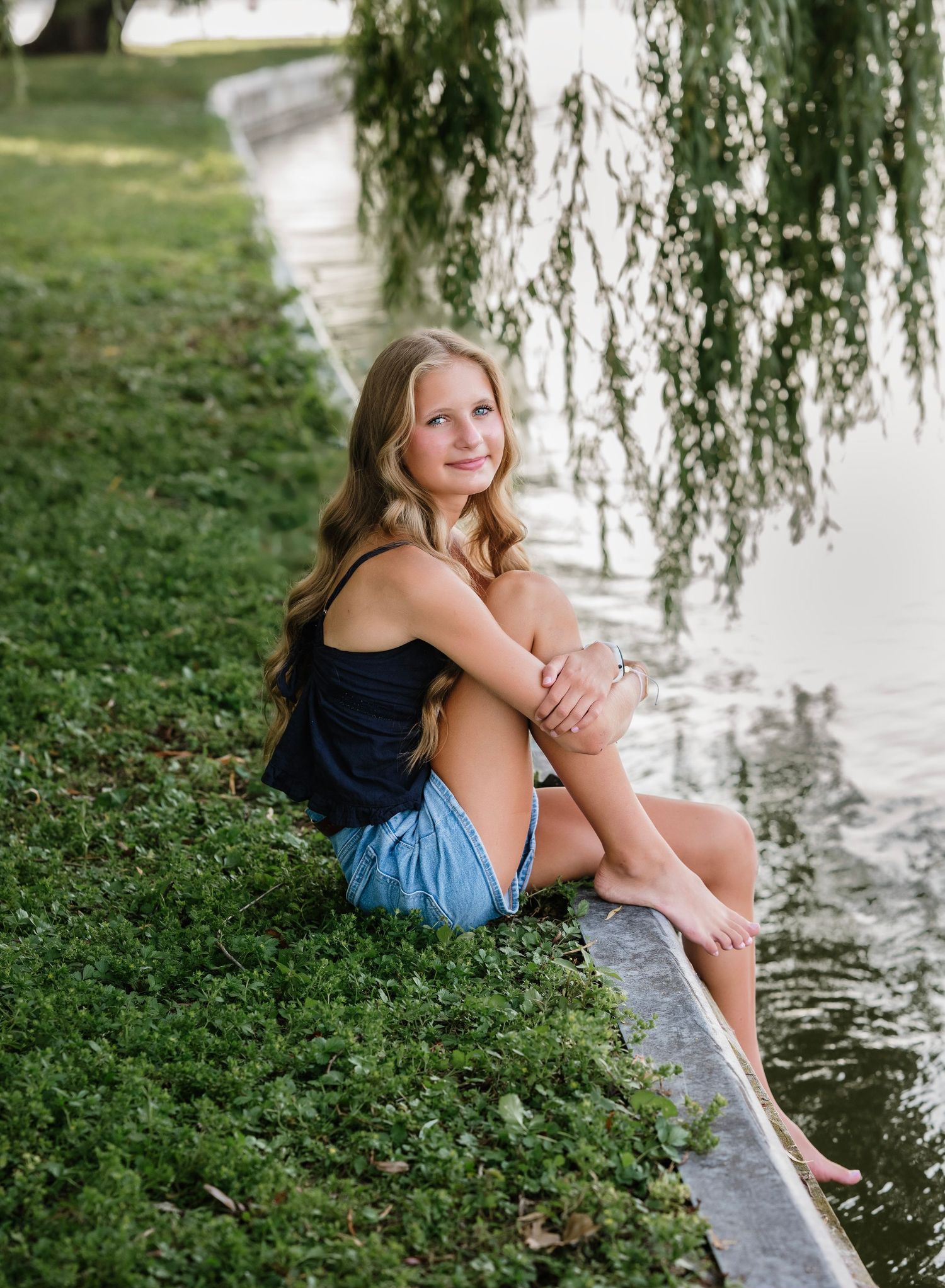 A person sits on grassy riverbank beneath a weeping willow tree on a sunny summer day.