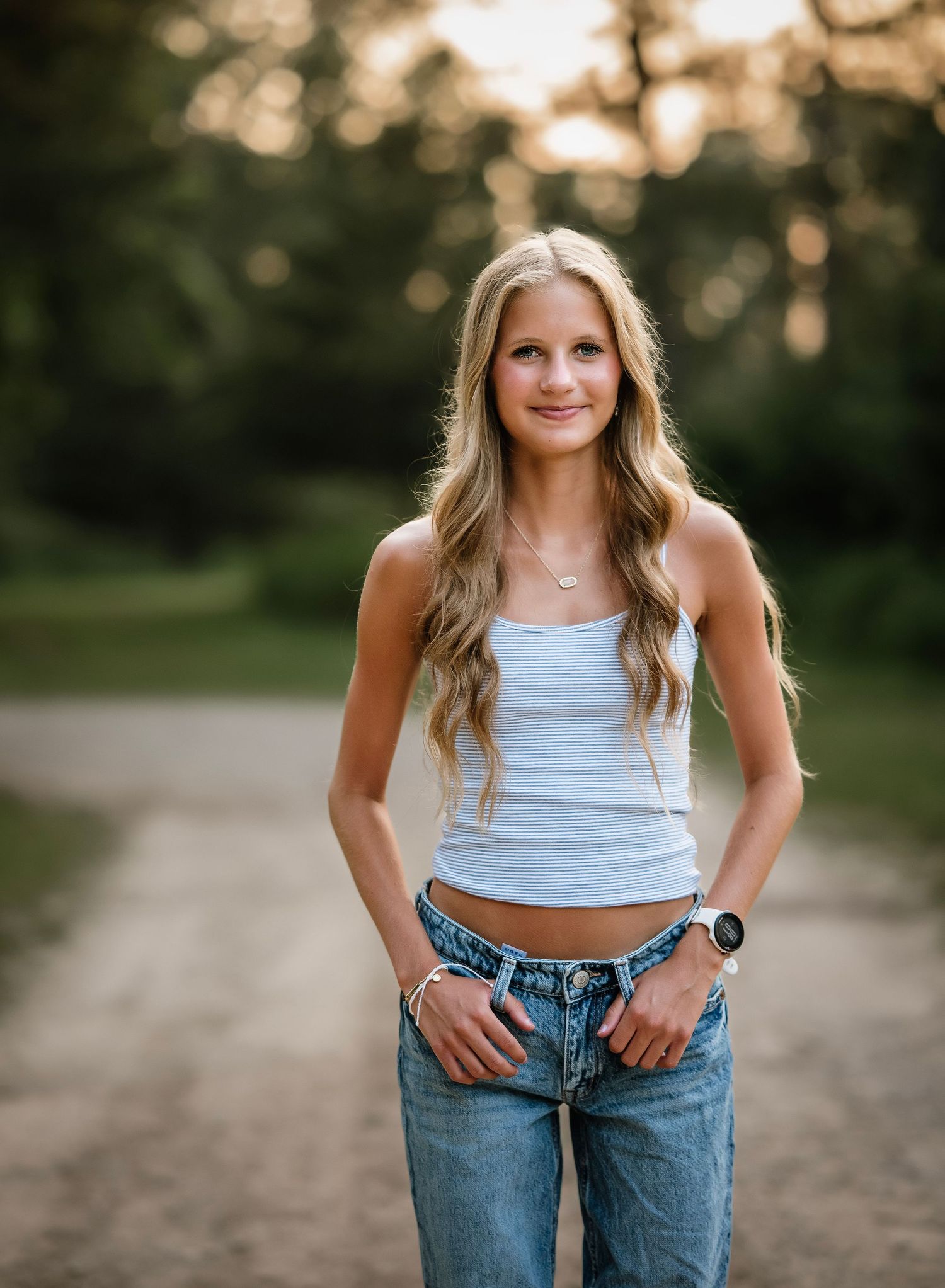 A person in a gray crop top and blue jeans poses casually outdoors during golden hour lighting.