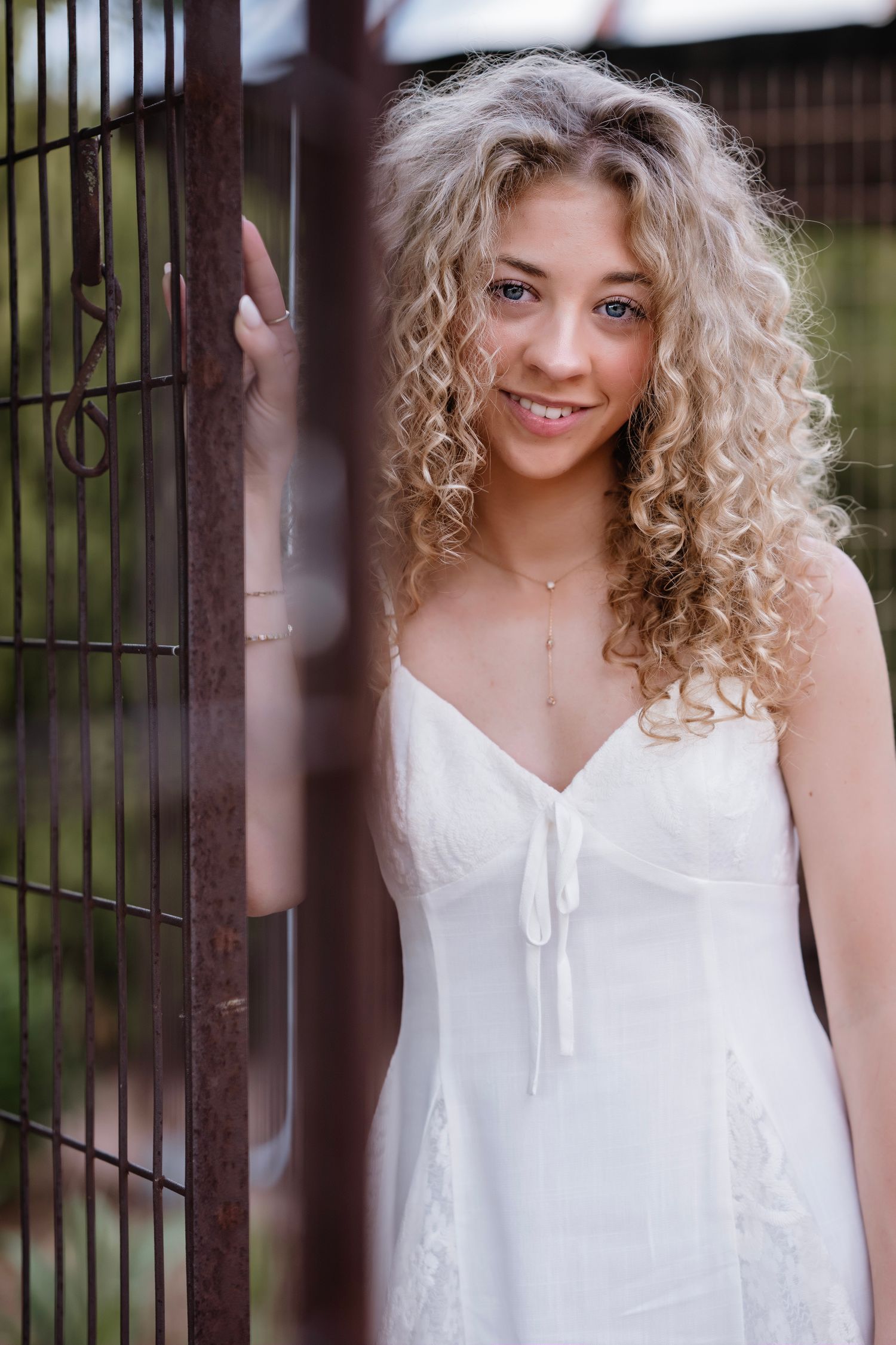 Young woman with curly blonde hair in white sundress smiling beside rustic metal gate outdoors.