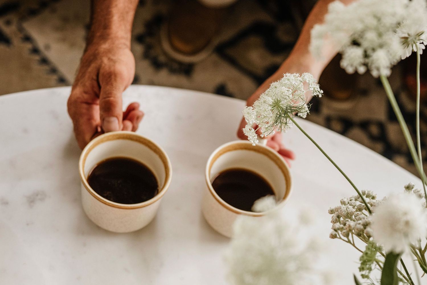Two hands placing ceramic cups of black coffee on a marble table beside white wildflowers.