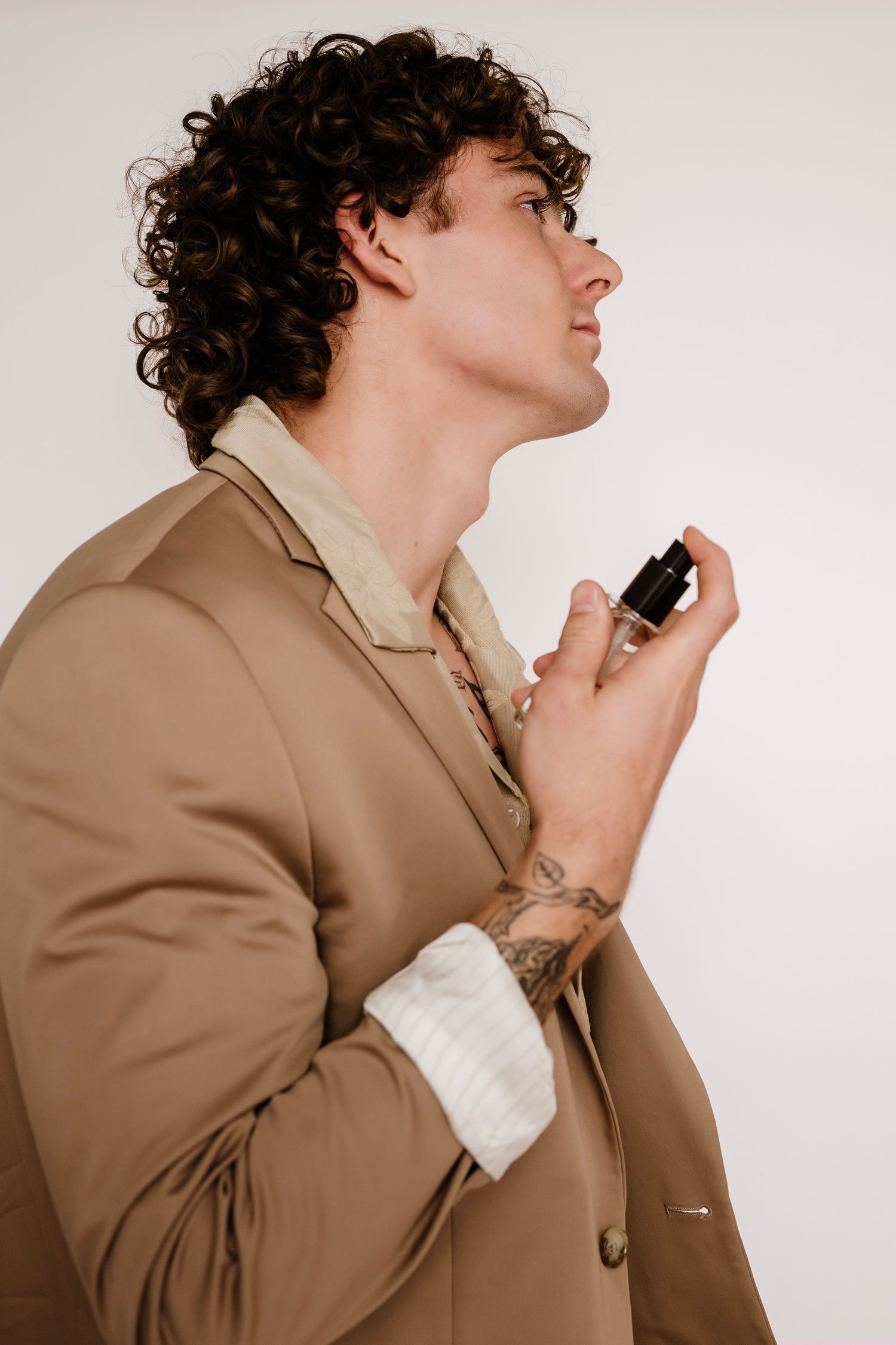 Man with curly hair in beige blazer applying cologne, side profile view, tattoo visible on wrist.