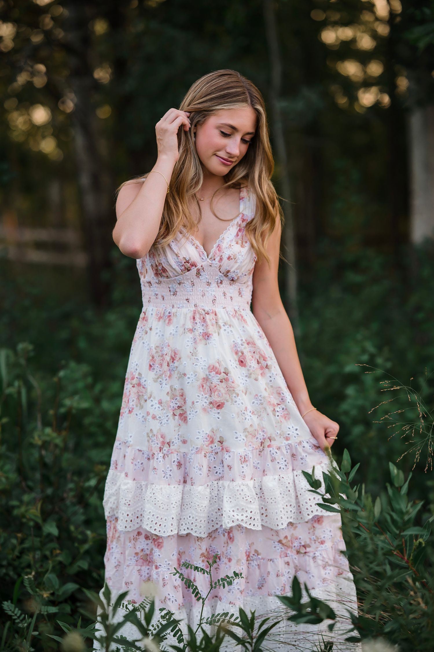 Young woman in a white floral tiered midi dress standing outdoors among lush green foliage in soft evening light.