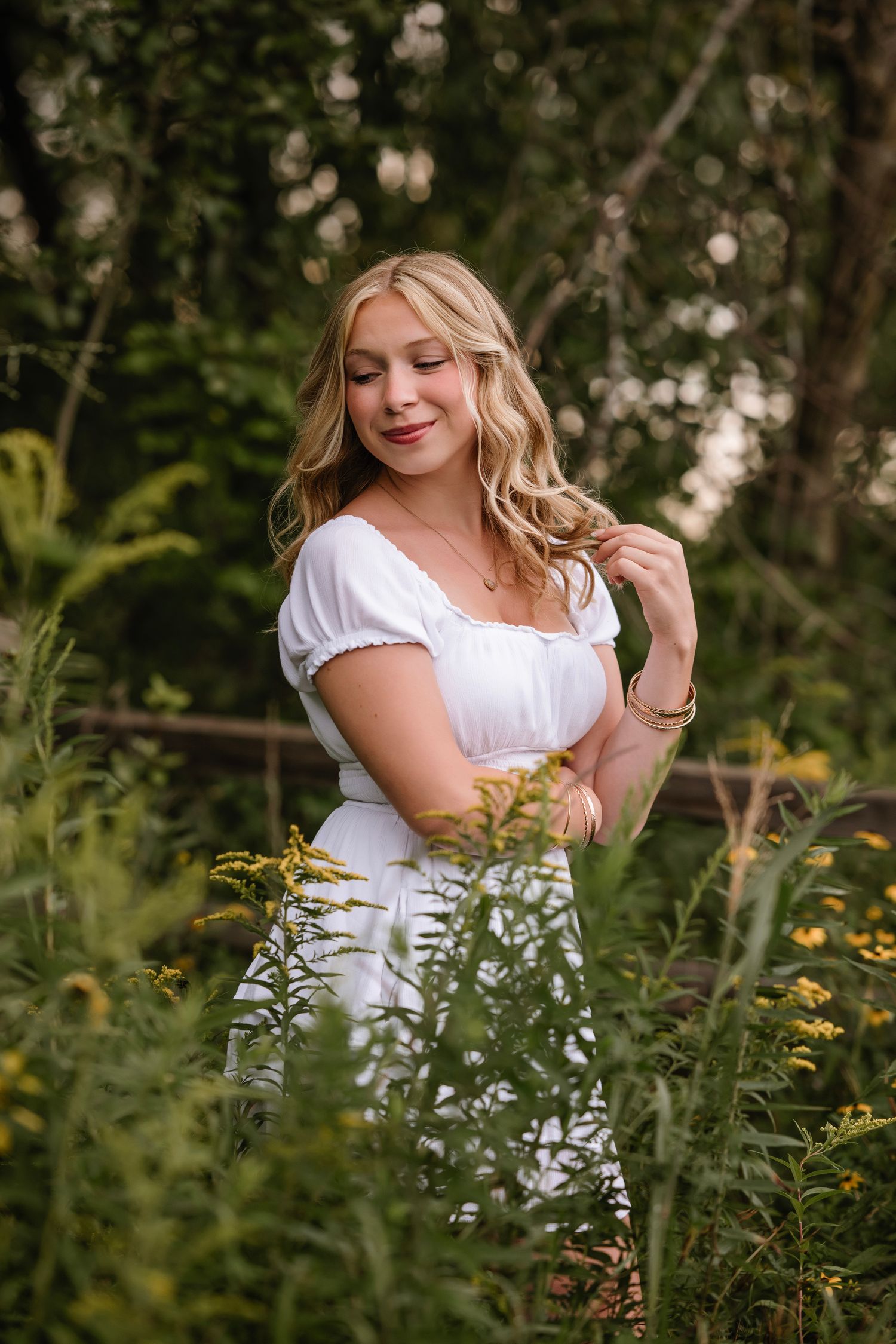 Young blonde woman in white dress poses among wildflowers and lush greenery in a serene outdoor setting.