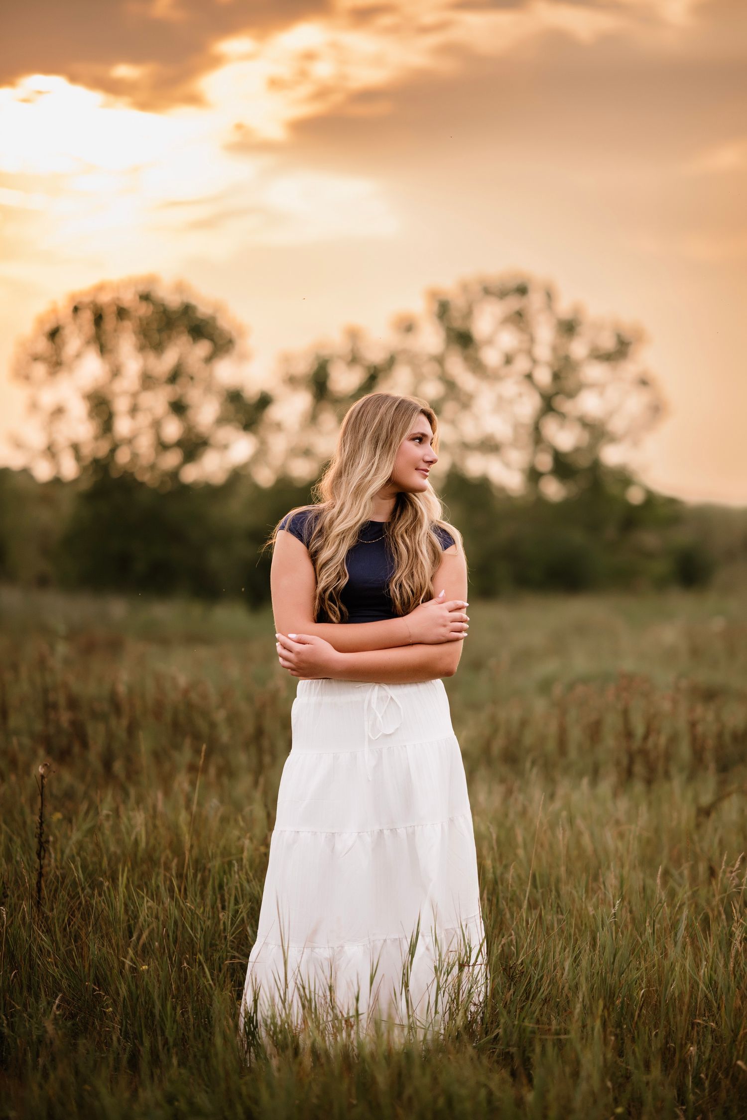 Young woman in white maxi skirt and navy top stands in golden field at sunset with arms crossed.