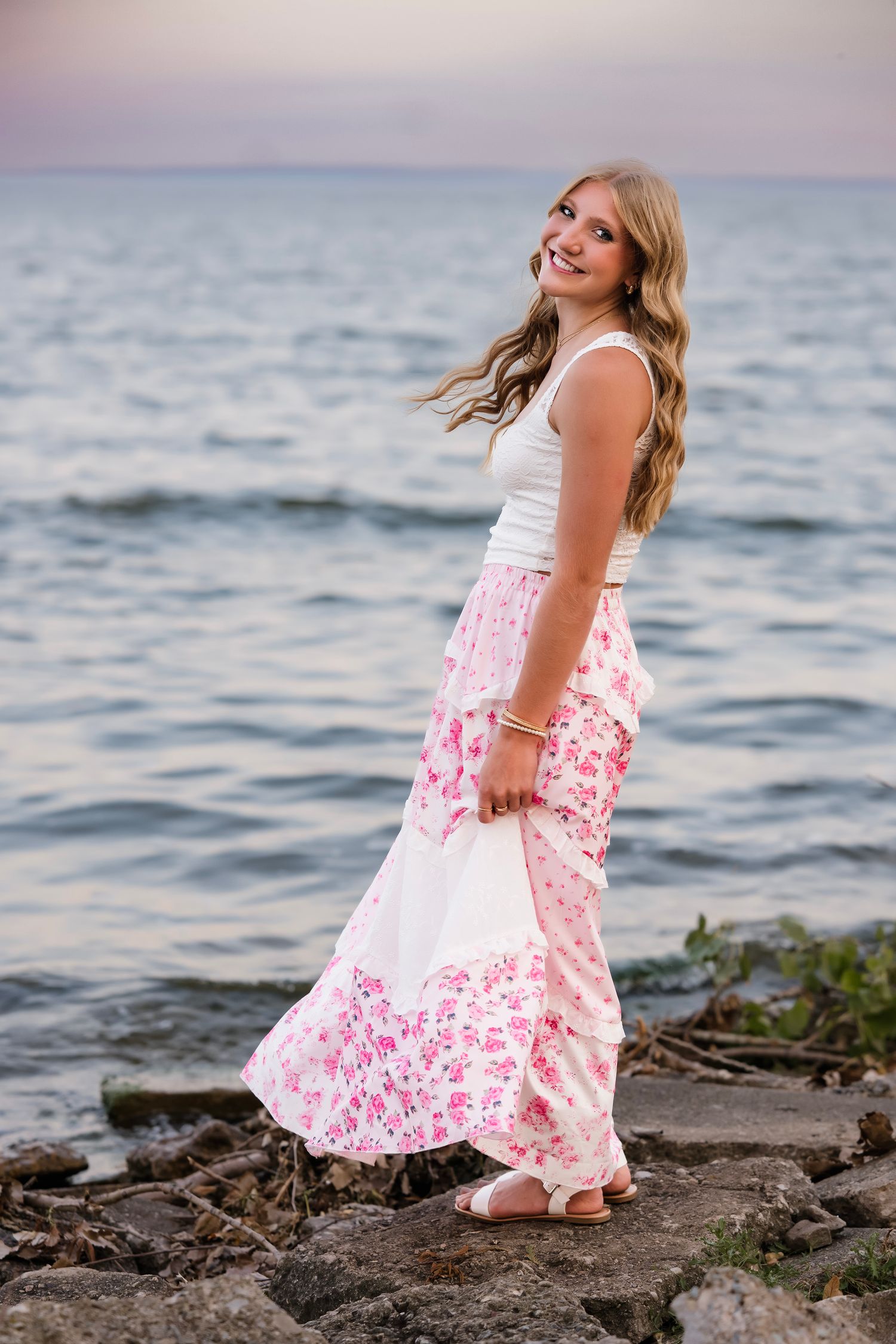 Smiling blonde teen in floral maxi skirt and white top poses on rocky shoreline at sunset.