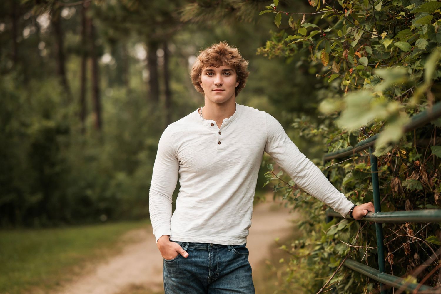 A person in a white henley shirt and jeans poses casually outdoors against a natural wooded background.