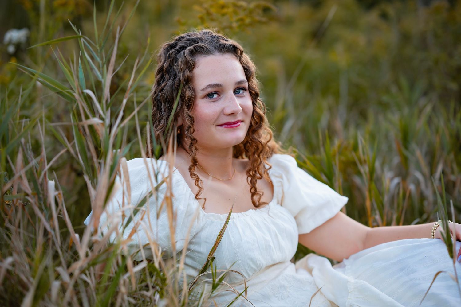 Young woman with curly hair in white dress sitting peacefully among tall grass in a natural outdoor setting.