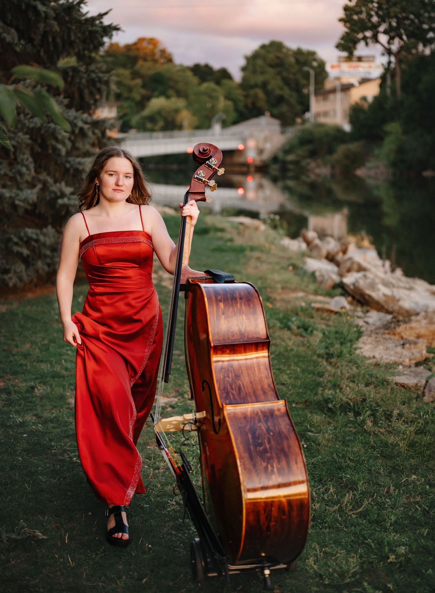 A high school senior in a flowing red dress poses with a double bass instrument against a scenic outdoor backdrop at sunset in Appleton.