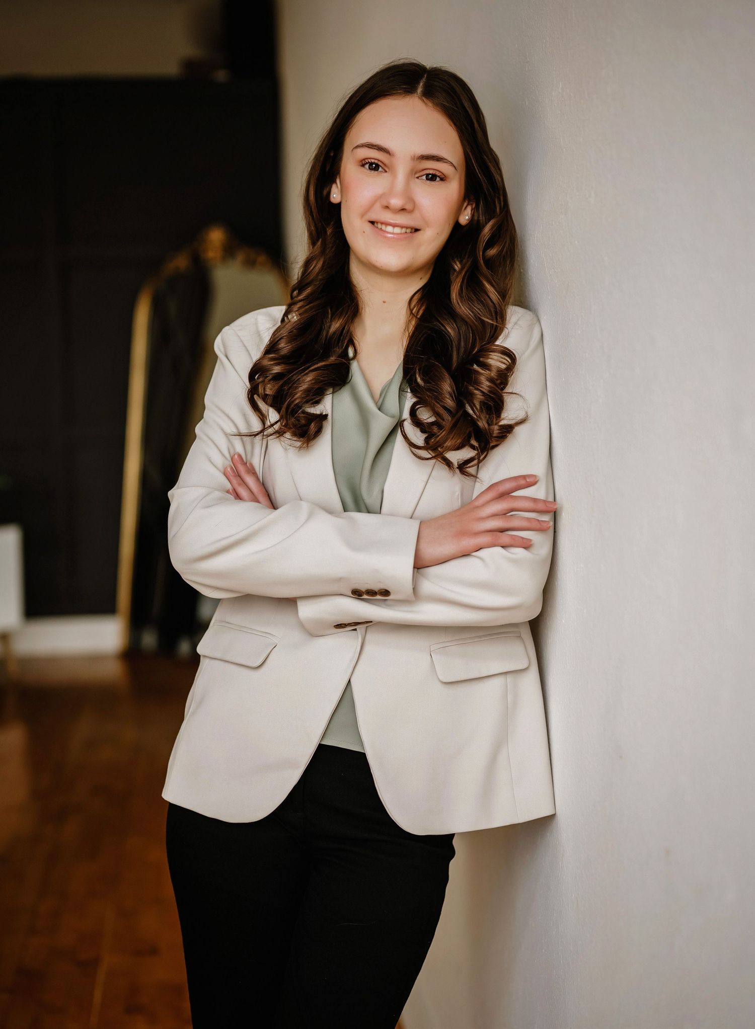 Professional portrait of someone wearing a cream blazer with green blouse and dark pants standing with crossed arms.