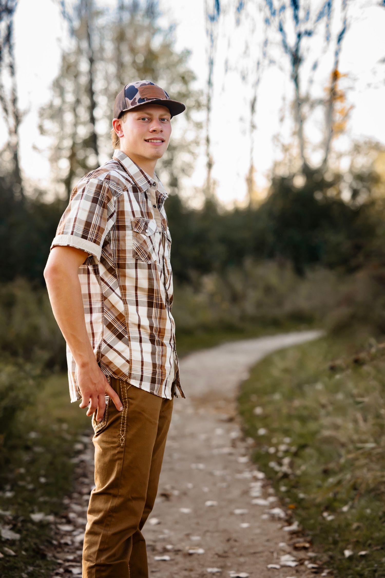 A person in a plaid shirt and brown pants poses on a dirt path through autumn woods at sunset.