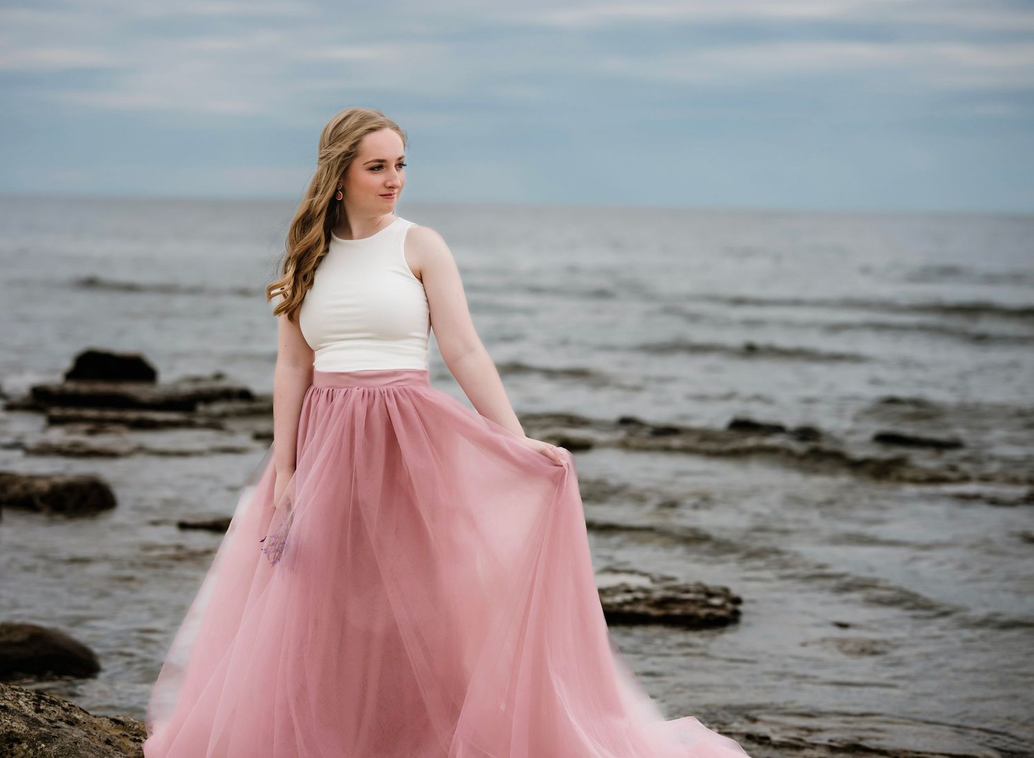 A person in a white crop top and flowing pink tulle skirt poses gracefully on a rocky beach at sunset.
