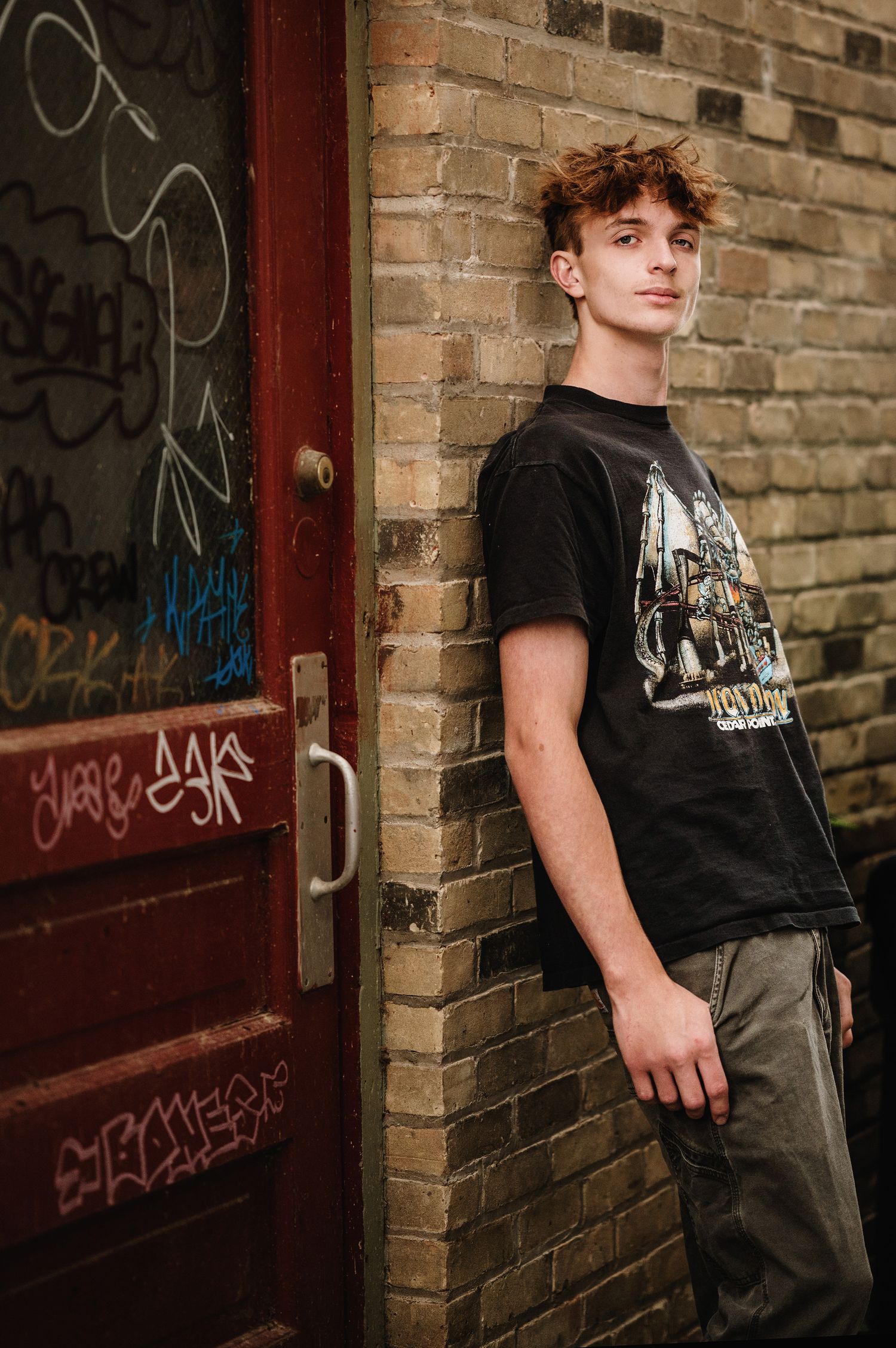 Person wearing black graphic t-shirt and gray pants poses against brick wall with graffiti in urban setting.