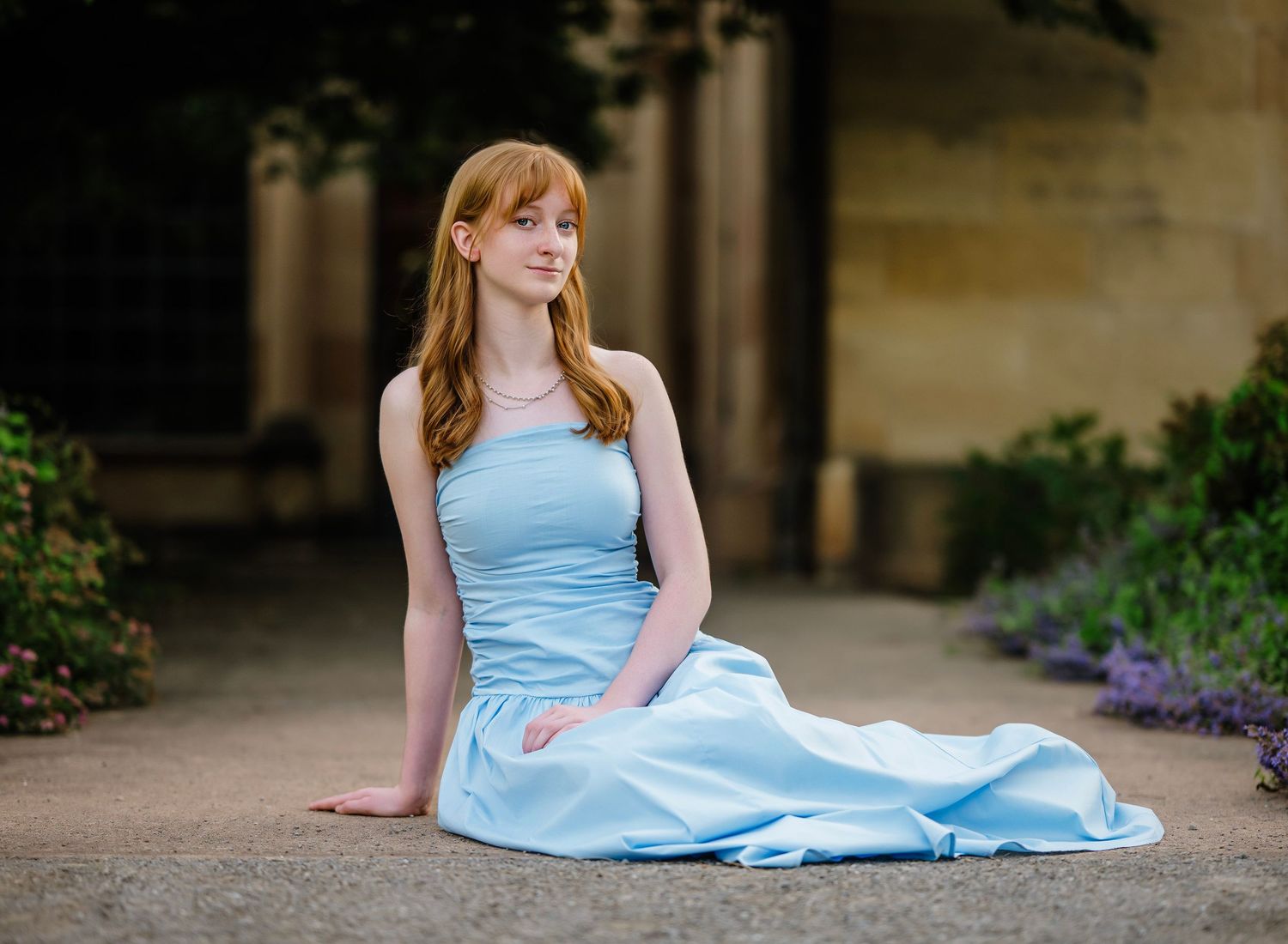 A person in a light blue dress sits gracefully on stone steps near blooming purple flowers in a garden setting.