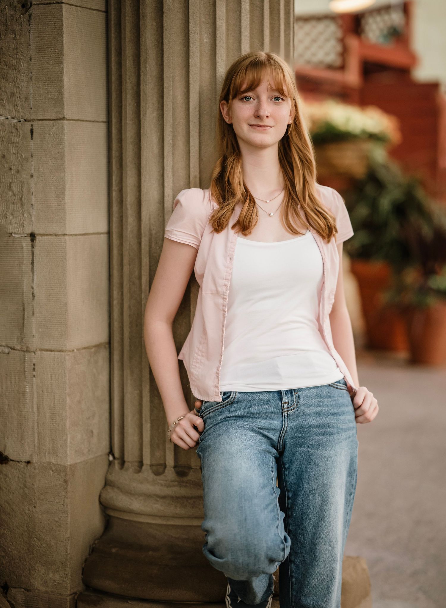 Person in pink shirt and jeans poses casually against stone columns in an outdoor setting.