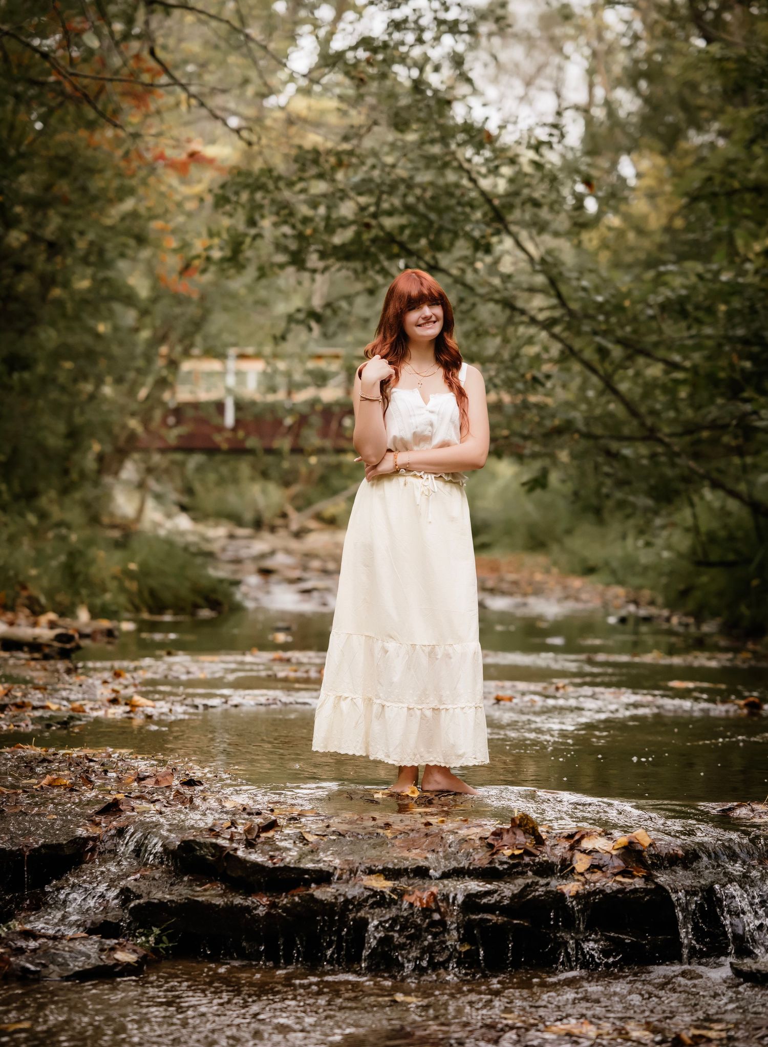 A person in a white lace dress stands on rocks by a flowing stream in a wooded setting during summer.